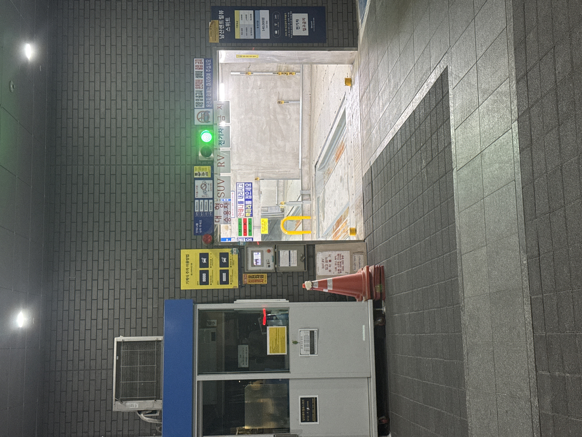 This image shows the entrance to an automated parking facility, presumably located at the Namsan Central View Suite in Seoul, South Korea. The scene is composed of a parking booth with an air conditioning unit attached. The booth has posted notices and instructions on how to use the automated parking system. A traffic cone is placed next to the booth. There are several signs attached to the brick wall, including parking rate information, instructions on how to use the automated parking system, and warnings prohibiting certain types of vehicles (SUVs, RVs, and electric vehicles) from entering. A green light indicates entry is permissible. The entrance to the parking structure is visible through the opening.