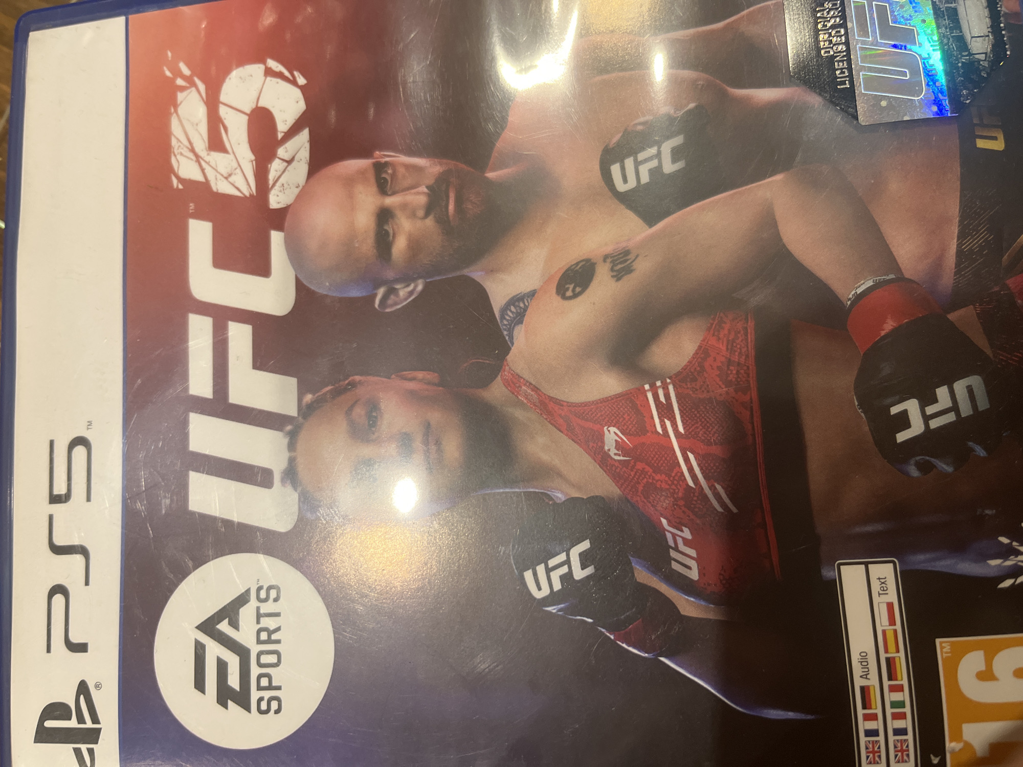 The image shows a copy of the video game "EA Sports UFC 5" for PlayStation 5. The cover art features two UFC fighters in action. The game case has a holographic UFC logo and a sticker indicating supported audio and text languages. A "16" rating symbol is also visible.