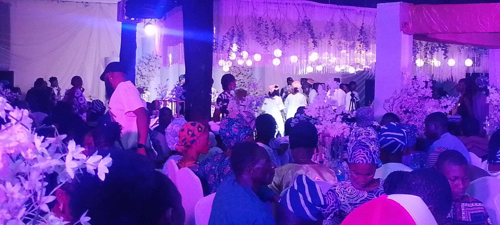 The image depicts an event occurring in Lokoja, Nigeria. Many people are present, with some seated at tables. The decorations include white flowers and strings of lights, possibly indicative of a wedding or formal celebration. In the background, several people are standing, while most of the foreground is filled with seated guests. Some are wearing traditional Nigerian attire, including headwraps. There is a stage area with more lights in the background. The room is lit with a purple hue.