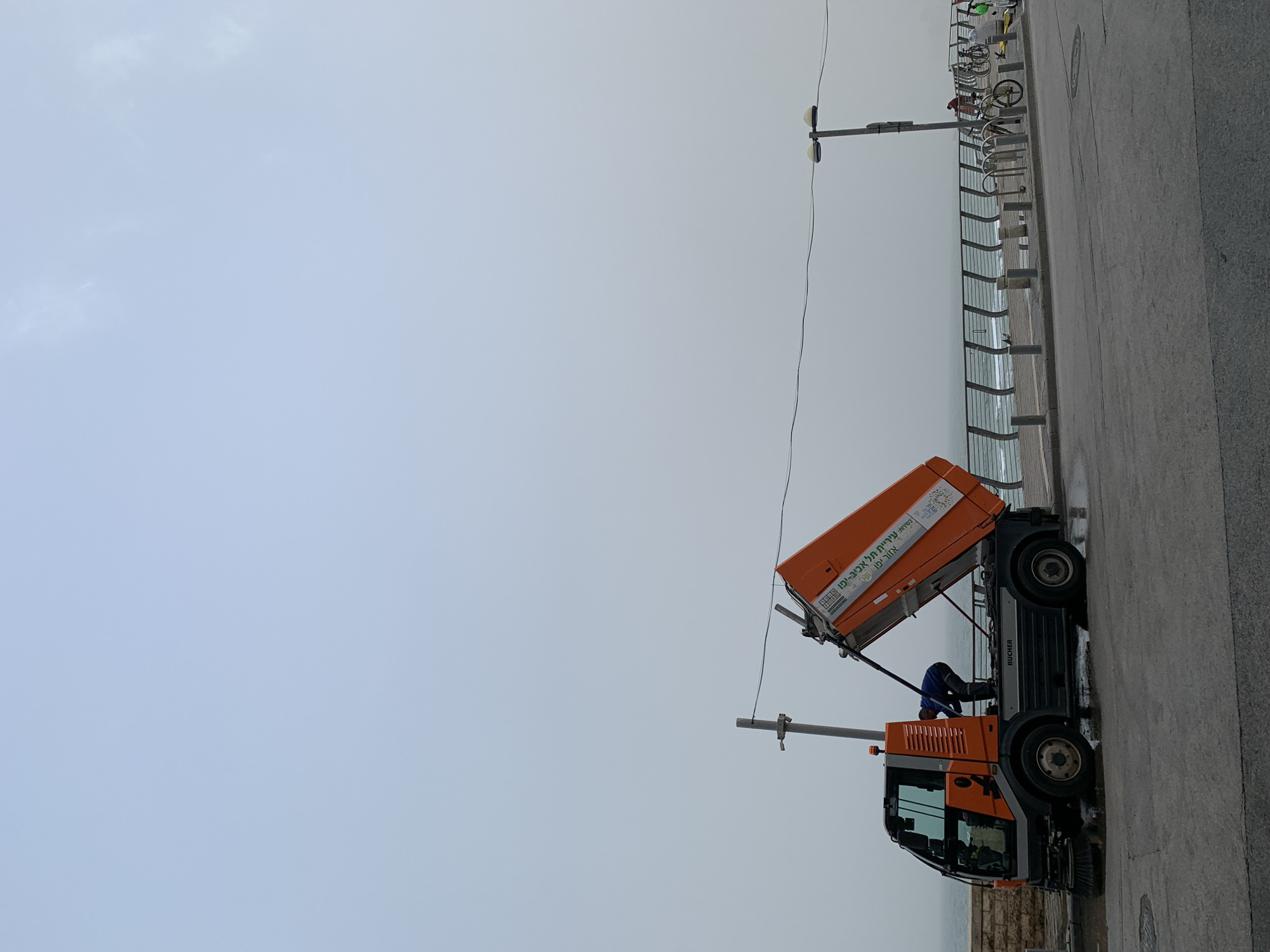 The image shows a street cleaning vehicle parked on a sidewalk alongside the ocean in Tel-Aviv, Israel. The truck, predominantly orange and black, has its rear container tilted upwards, presumably in the process of emptying collected waste. An individual wearing a blue shirt is bent over, likely attending to the vehicle's operation.
A metal railing separates the sidewalk from the beach and the ocean, partially obscured by fog or mist. A street lamp post is visible near the vehicle, and a bicycle rack filled with bikes is further down the sidewalk. The sky is overcast, suggesting cloudy weather conditions. The scene appears to be a coastal promenade where routine maintenance or cleaning is taking place. The truck is branded with the words "תל אביב -יפו" (Tel Aviv-Yafo) confirming the location.