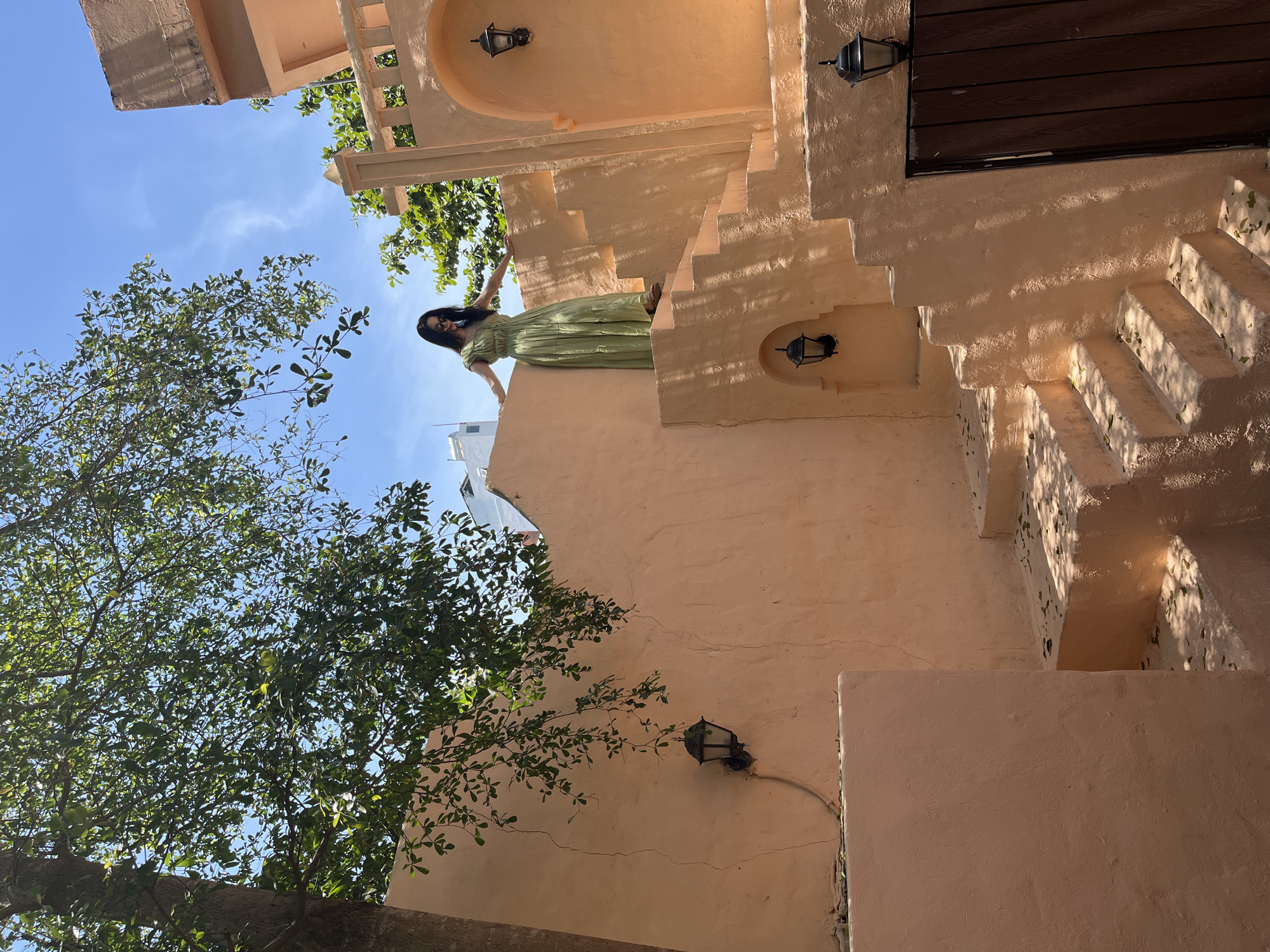 A low-angle view captures a woman standing on a peach-colored, textured building with stairs, possibly in Đà Nẵng, Vietnam. The architecture features rounded arches and decorative lighting fixtures. The woman is wearing a green dress and sunglasses. Lush green trees are visible to the left, partially obscuring the sky. The lighting casts strong shadows, adding depth to the scene.