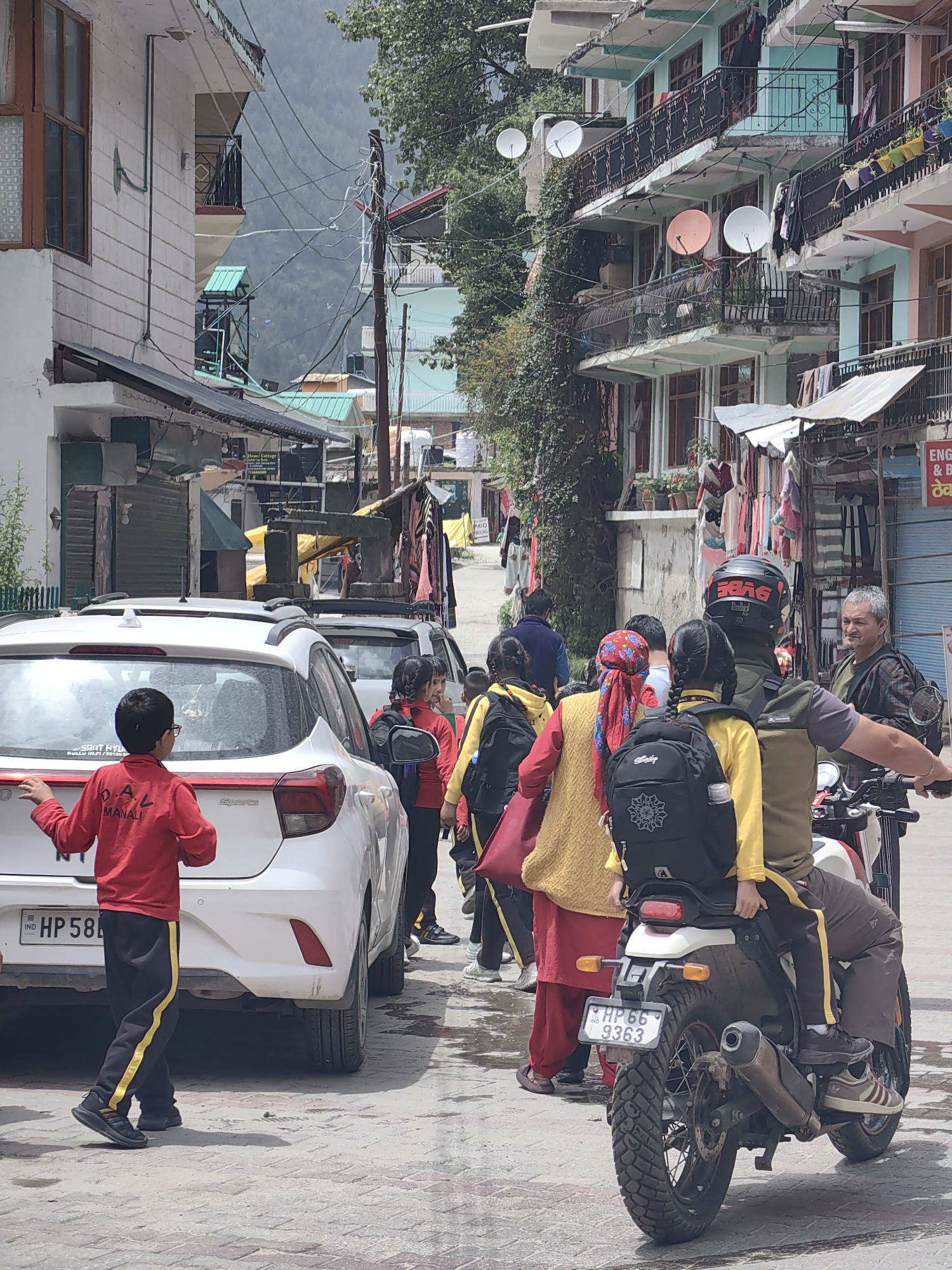 The image depicts a street scene in Vashisht, India, likely a busy commercial area with buildings lining both sides of a narrow road. In the foreground, a young boy in a red school uniform with black and yellow striped pants stands next to the rear of a white car, possibly a Hyundai i20, with the license plate "HP 58 M". To his right, a group of people, predominantly children in similar school uniforms, are walking or standing. One girl in the group has a red headscarf.
Behind the children, a man wearing a black helmet is riding a motorcycle, with another child seated behind him. The child on the motorcycle is wearing a yellow top and dark pants. The motorcycle's license plate reads "HP 66 9363". To the right of the motorcycle, an adult man with graying hair, wearing a plaid shirt, looks towards the group.
The buildings in the background are multi-story structures with balconies, some decorated with potted plants and satellite dishes. Clothes are visible hanging from balconies, suggesting residential or mixed-use buildings. The street itself is paved with cobblestones or similar material. The overall atmosphere appears to be one of daily life and movement within a densely populated area. The presence of school children and various vehicles indicates an active community.