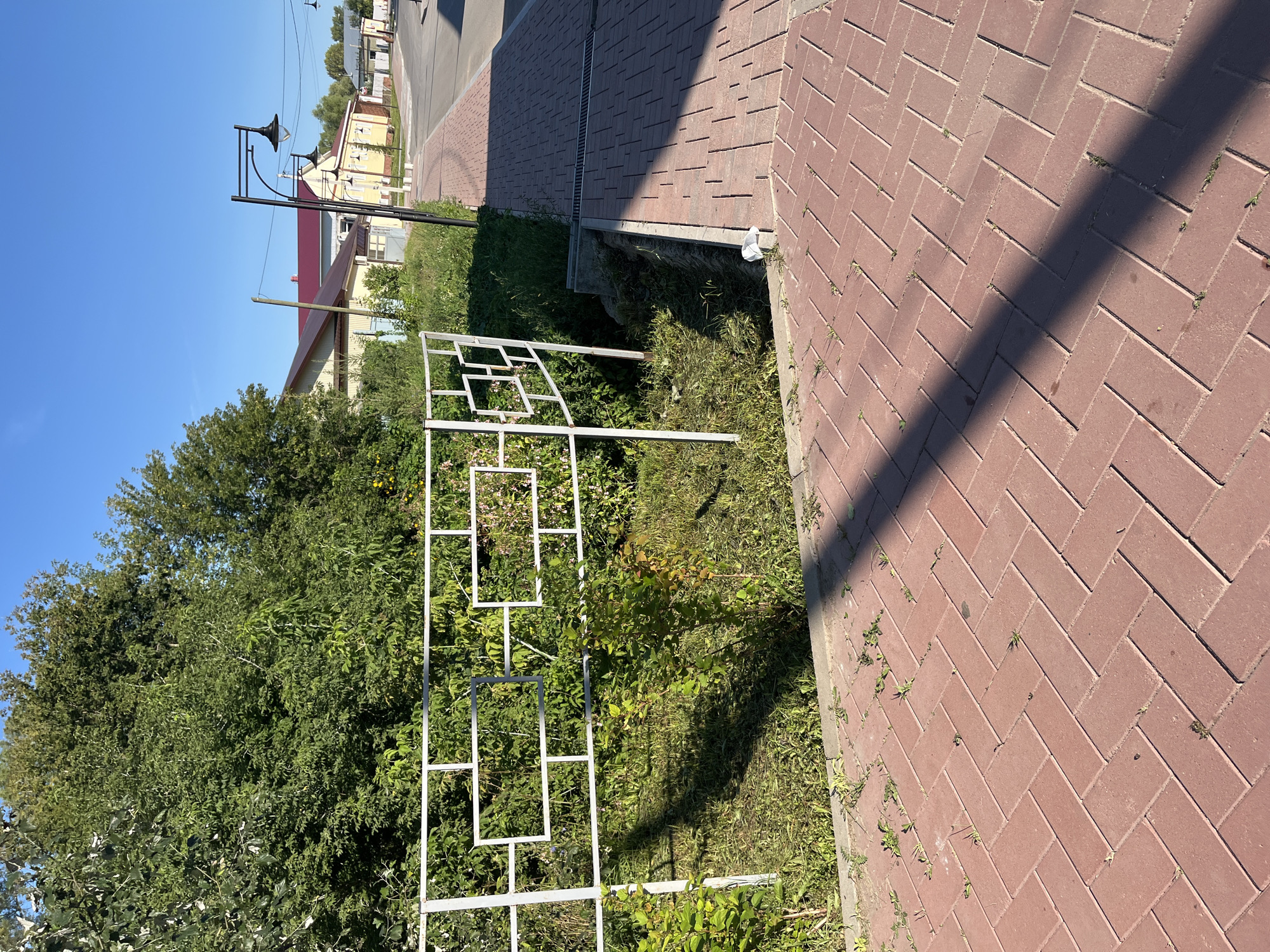 The image depicts a sunny day in Blagoveshchensk, Russia, showcasing a section of a sidewalk and adjacent greenery. The sidewalk is paved with red brick pavers laid in a herringbone pattern. A white metal fence with geometric patterns runs along the edge of the sidewalk, separating it from a patch of overgrown grass and bushes. Beyond the fence, trees with lush green foliage dominate the landscape, with some smaller bushes and flowers visible. In the background, a street curves to the right, lined with yellow buildings with red roofs and several lampposts. The sky is a clear, vibrant blue, indicating pleasant weather conditions. No people are visible in the frame, and the scene appears to be a typical urban or suburban streetscape.