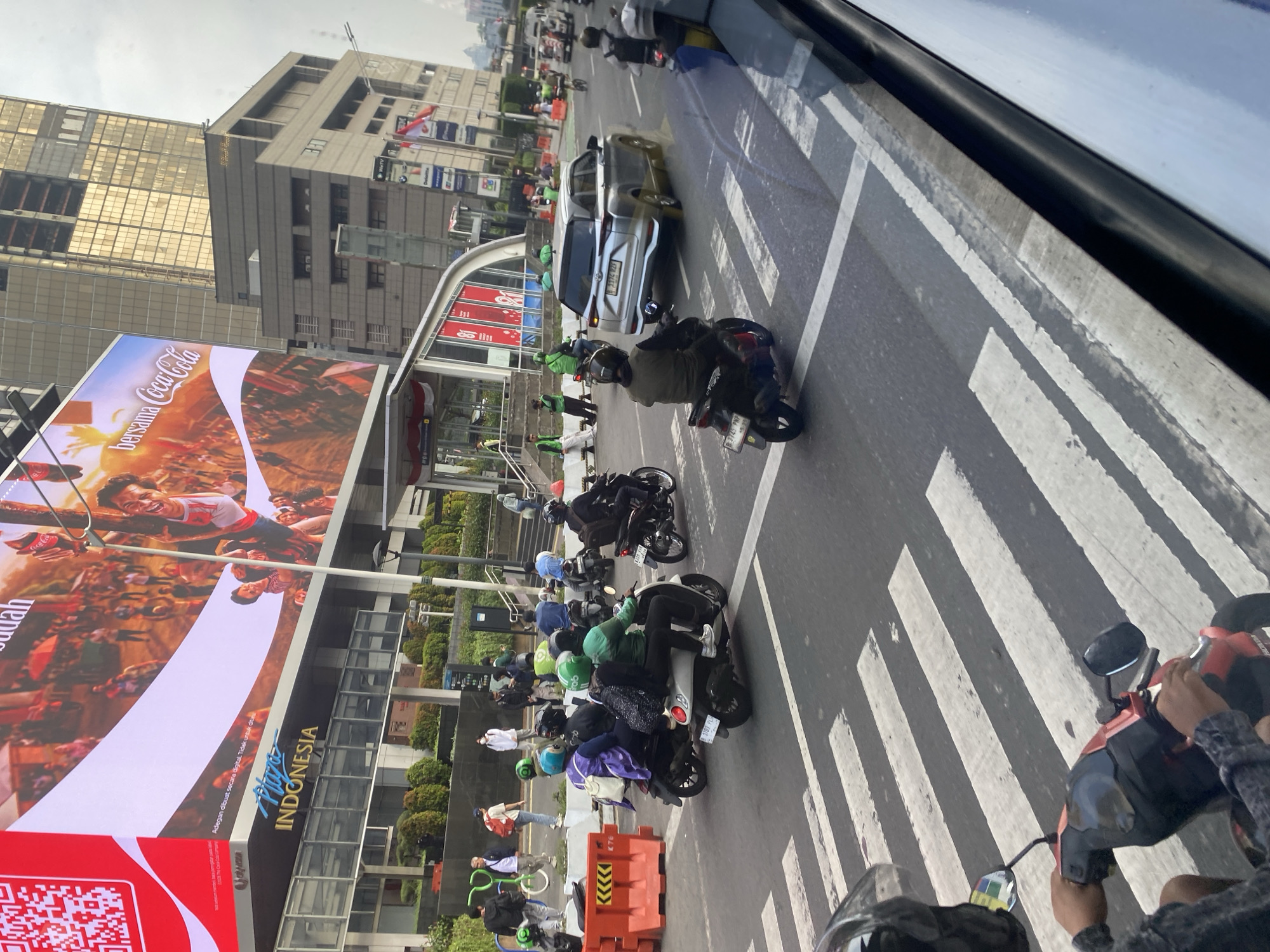 The image depicts a street scene in Central Jakarta, Indonesia, likely in the vicinity of Plaza Indonesia, as indicated by the signage. In the foreground, a person is riding a red motorcycle, seen from a first-person perspective, crossing a pedestrian zebra crossing. The road is paved and has white stripes marking the pedestrian crossing. Several other motorcycles are visible on the road, some carrying passengers, and a silver SUV is also present. A prominent billboard displaying an advertisement for Coca-Cola is visible in the background, along with modern buildings and lush green landscaping. The sky is overcast. People are seen walking and riding motorcycles on the sidewalk and road, indicating a busy urban environment. Orange traffic barriers are also placed on the side of the road.