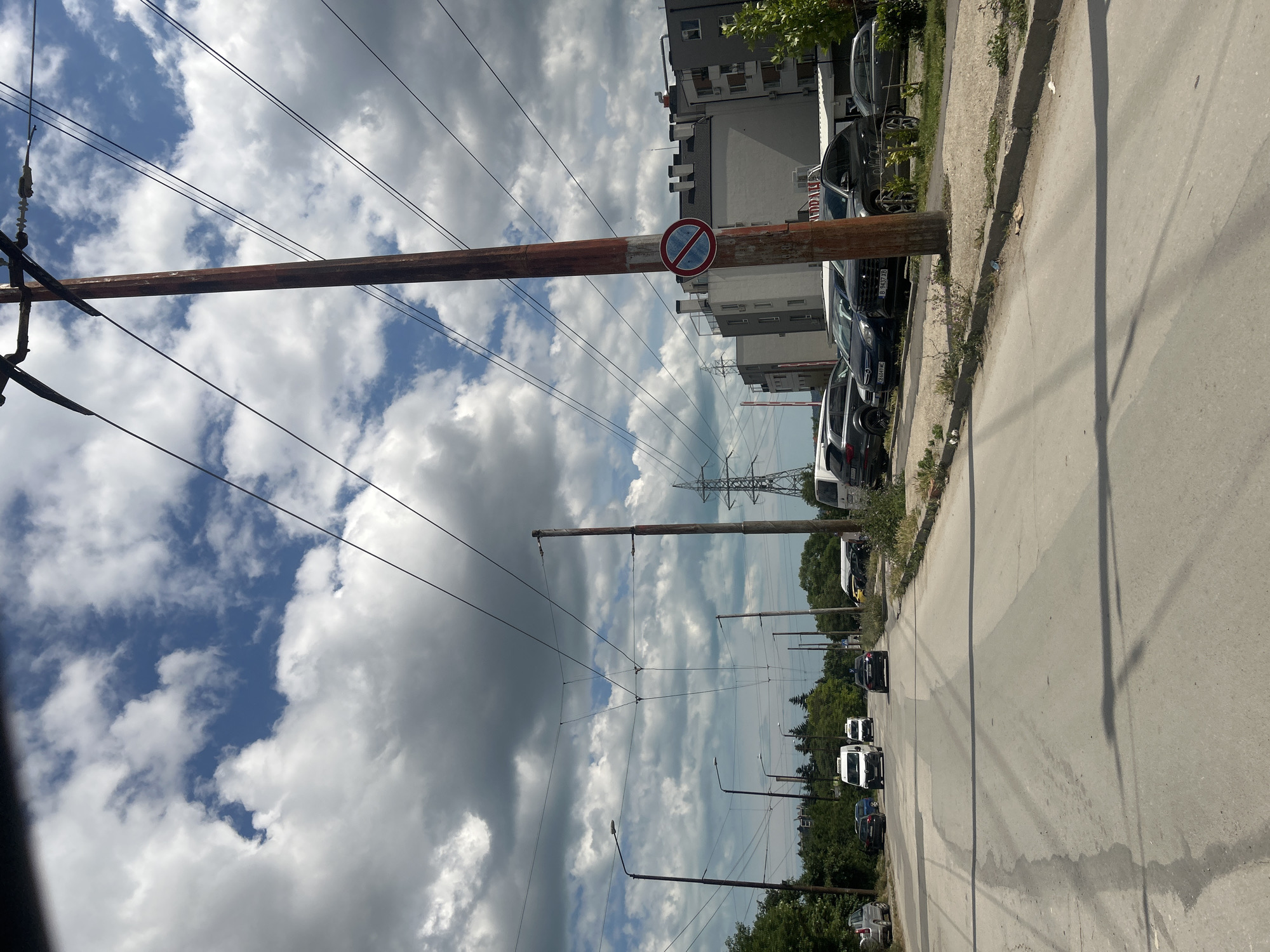 The image depicts a street scene in Varna, Bulgaria, under a cloudy sky. The view is from a low angle, possibly from a vehicle, looking down a paved road lined with trees on the left and buildings and parked cars on the right. Overhead, numerous electrical wires crisscross the sky, supported by wooden and metal poles. One pole in the foreground on the right has a circular red and blue sign with a diagonal line through it, indicating a prohibition. Several vehicles, including vans and cars, are visible on the road and parked along the side. The environment appears to be urban or suburban, with a mix of residential and commercial buildings. The overall lighting suggests it is daytime.