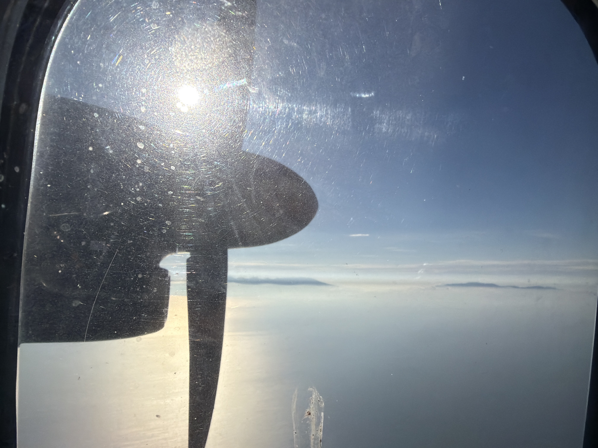 An aerial view from an aircraft window shows a propeller blade in the foreground, casting a shadow. The sun is visible and flares strongly through the window. Beyond the window, the sky is a clear blue, meeting a layer of clouds. Below the clouds, a body of water stretches to the horizon, with islands visible in the distance, suggesting a location near Yuzhno-Kurilsk, Russia. The windowpane is smudged and scratched, with visible dust particles.