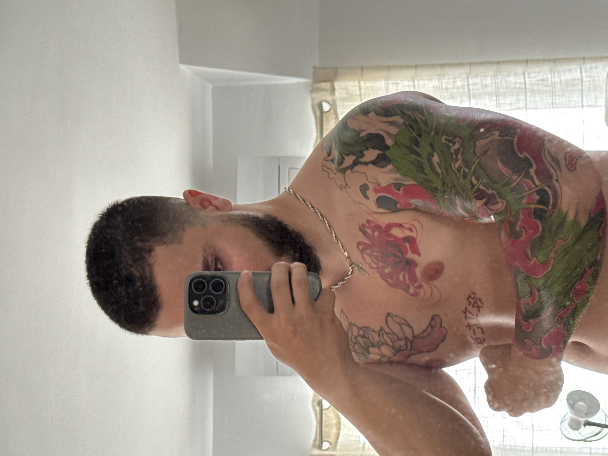 A man is taking a mirror selfie. He has short, dark hair, a beard, and is wearing a gold chain. The upper portion of his body is visible, and he has tattoos covering his shoulder, chest, and arm. He is holding a cell phone in his left hand, partially obscuring his face. The background includes a white wall and a curtain. There is nothing to indicate that the image was taken in Plovdiv, Bulgaria.