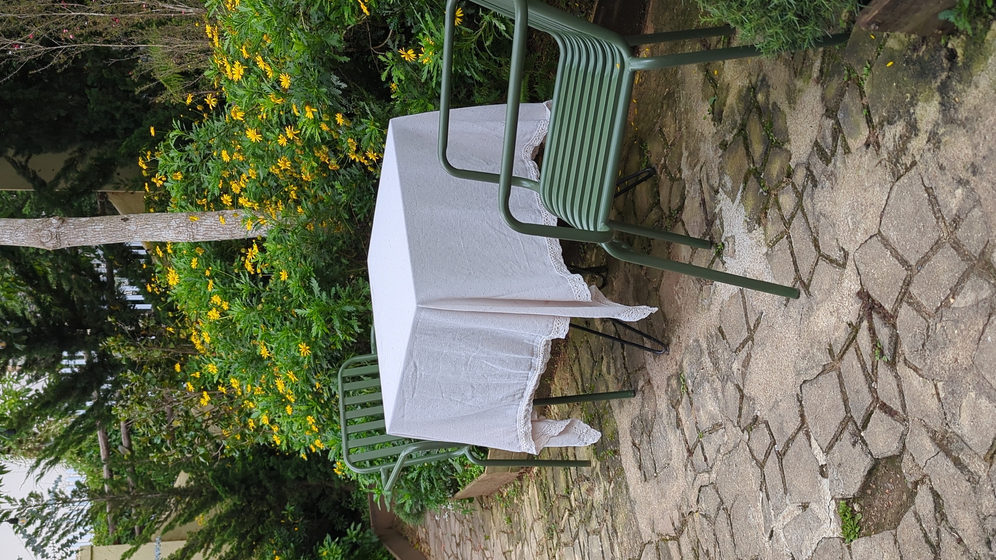 In the city of Lâm Viên - Đà Lạt, Vietnam, a charming outdoor seating area is depicted. A square table draped with a white tablecloth, adorned with delicate lace trim, sits centrally. Two green metal chairs, one on each side of the table, are positioned for seating. The setting is a garden with abundant greenery, featuring dense bushes with small yellow flowers and mature trees. The ground is paved with irregular grey stones, and some areas show patches of moss and weeds, suggesting a slightly weathered but well-maintained outdoor space. The overall impression is one of tranquility and natural beauty.