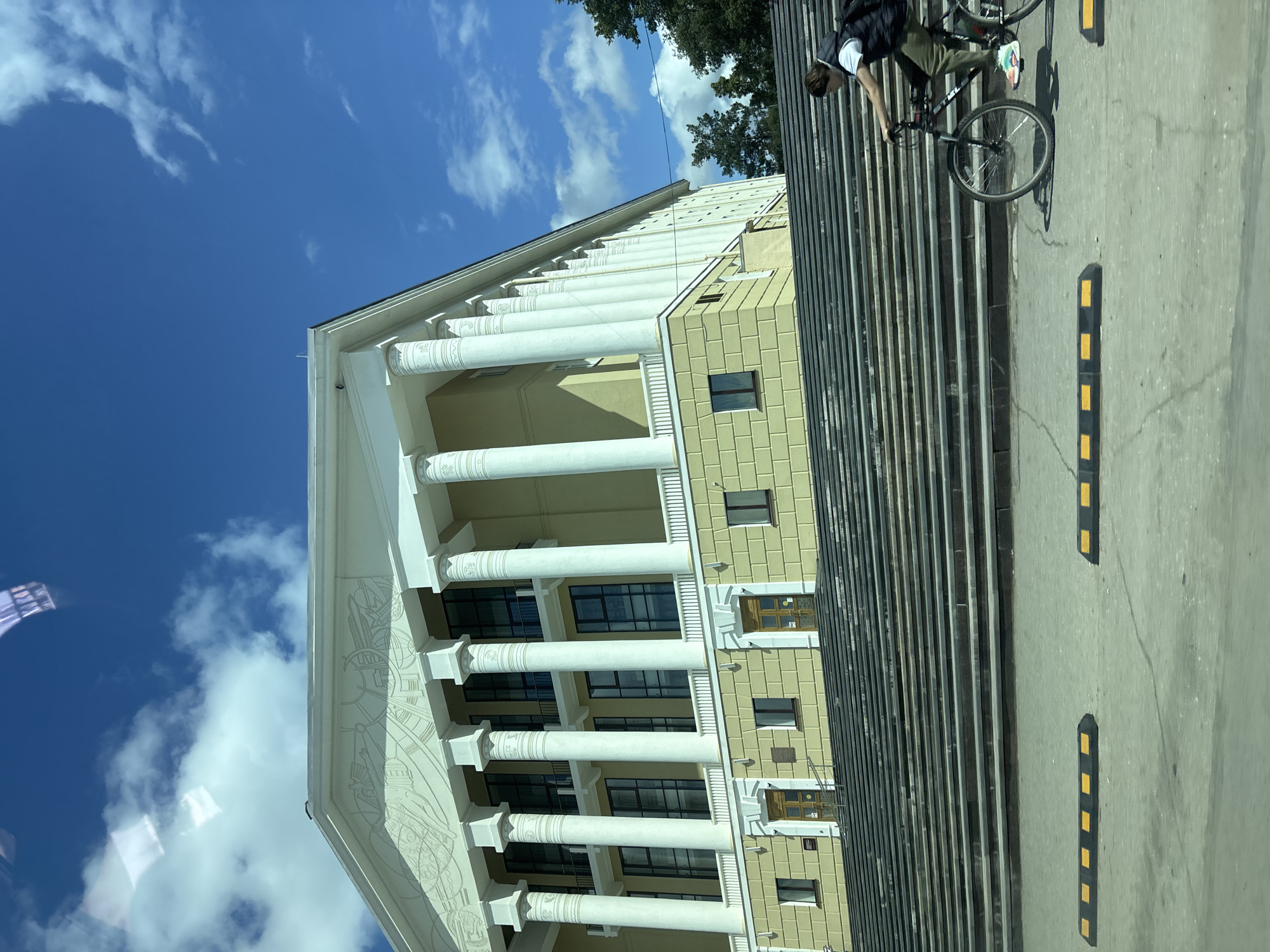 The image depicts a multi-story building with classical architectural elements, including a facade adorned with white columns. The building is light yellow with rectangular window patterns. Adjacent to the building is a set of dark-colored stairs.
A cyclist is on a bicycle on the stairs. The surrounding environment includes a partly cloudy blue sky.
Based on the architectural style and known landmarks, the building is likely the Votkinsk City Administration building in Votkinsk, Russia.