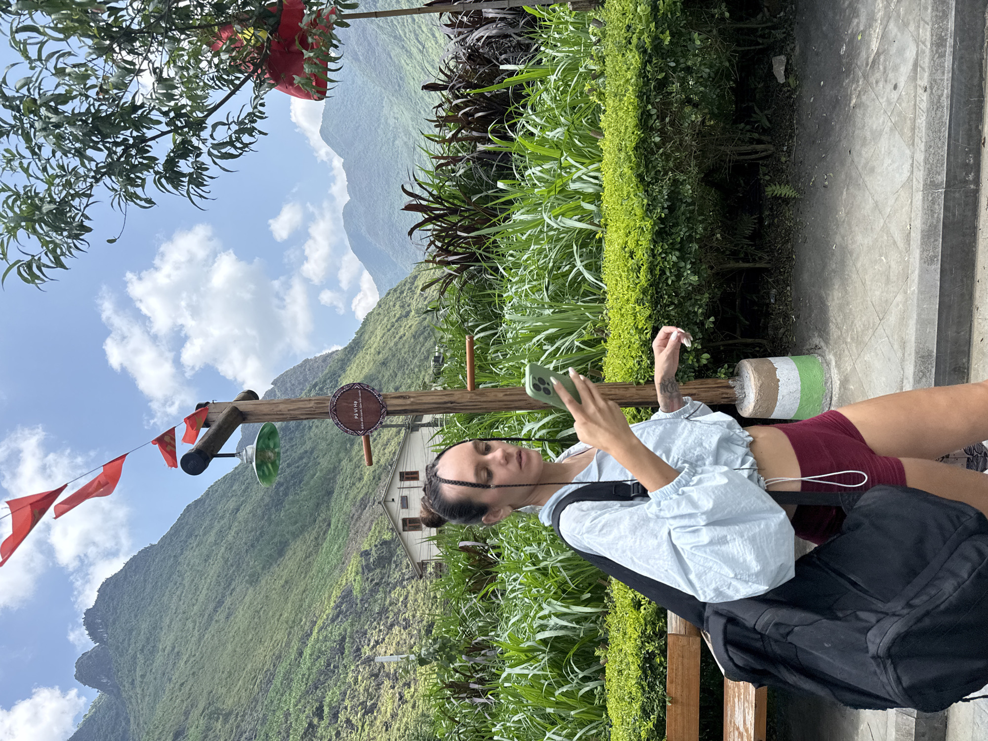 A young woman sits on a low bench, holding a cell phone, with a rural, mountainous landscape in the background. She has dark hair braided in front and a bun at the back, fair skin, and is wearing a light blue jacket and maroon shorts. A black backpack sits next to her on the bench. The bench is painted with green, white, and brown stripes. The background includes green, grassy hills and mountains under a blue sky with white clouds. There is also a wooden post with a sign and red flags, a two-story white house, and various plants and greenery. The location is Mèo Vạc Commune, Vietnam, as indicated on the sign post.