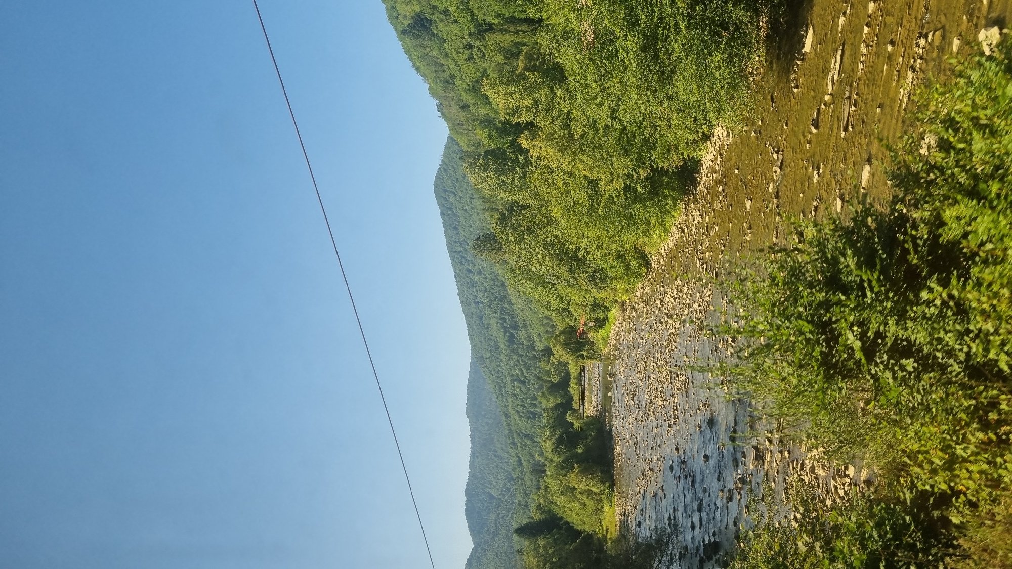 The image shows a scenic view of a clear, shallow river flowing through a valley. The riverbed is visible, covered with numerous small stones and pebbles. Lush green trees and vegetation line the banks of the river, leading up to rolling, tree-covered hills in the background. The sky is clear and blue. In the distance, a small wooden bridge spans the river, and a hint of a red-roofed building is visible among the trees. A thin black wire runs horizontally across the top portion of the image. This scene is located in the city of Skole, Ukraine, which is known for its natural beauty and mountainous terrain.