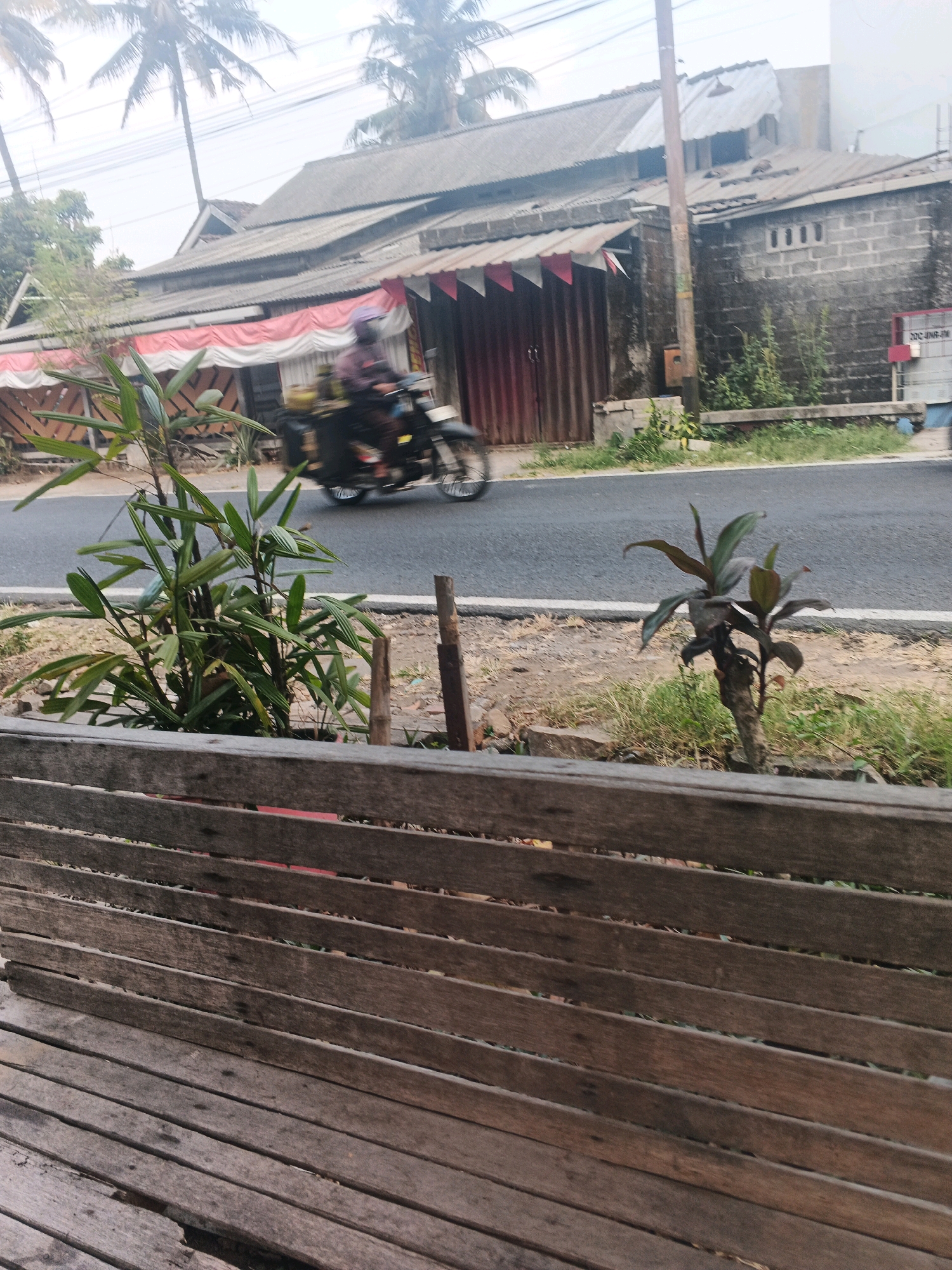 The image depicts a street scene in Ungaran, Indonesia. In the foreground, a wooden bench is visible, made of weathered planks. Beyond the bench, a paved road runs horizontally across the frame. On the road, a person is riding a motorcycle, their form blurred due to motion. To the left of the road, there is a plant with lush green leaves. Further back, buildings are situated along the roadside. One building features a roof made of corrugated metal and a facade decorated with red and white banners. Another adjacent building is constructed from grey bricks. Tall palm trees with fronds are visible in the background, suggesting a tropical or subtropical environment. The overall impression is of a residential or commercial street on a slightly overcast day.