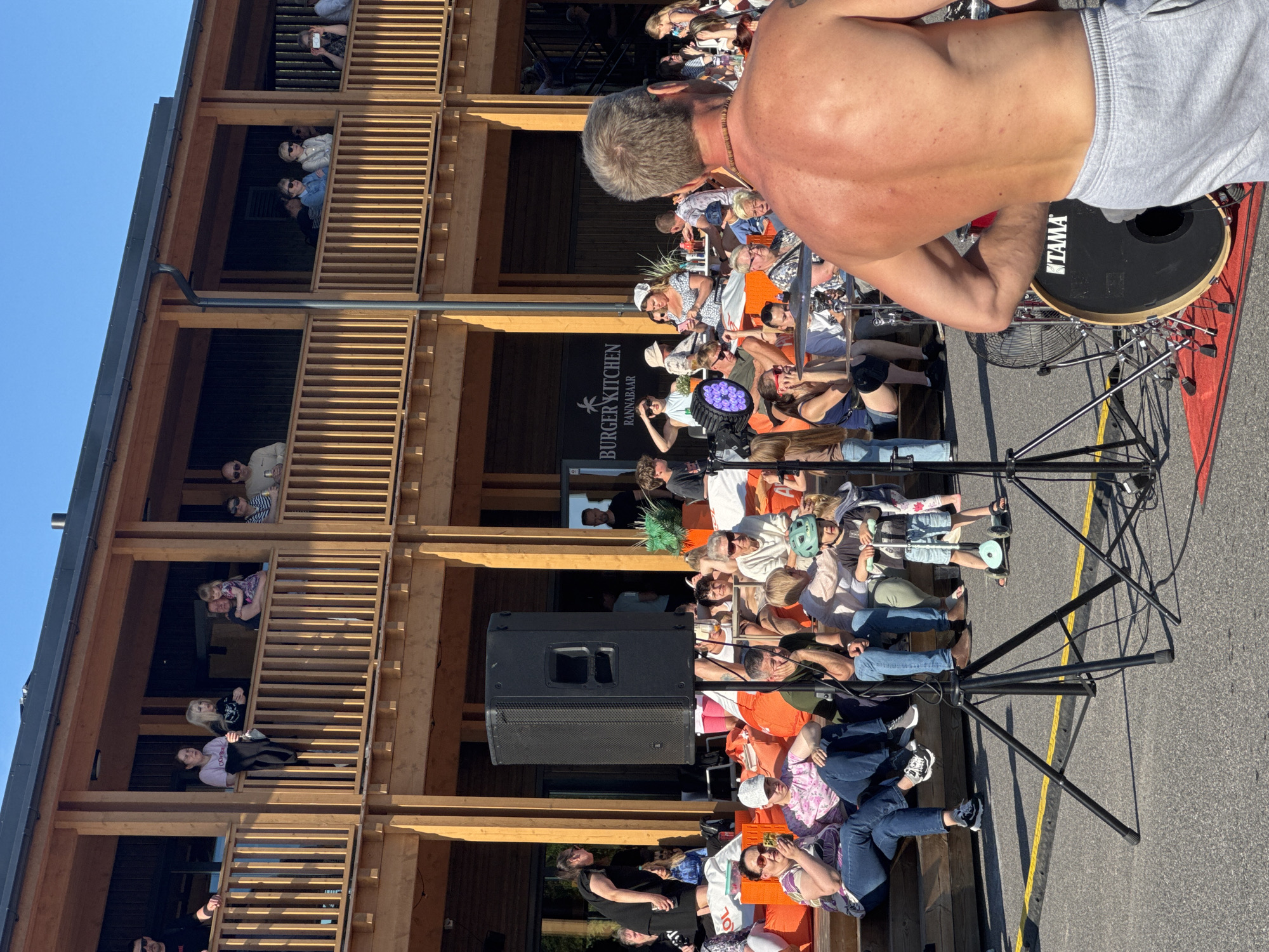 This is a vertical, low-angle, outdoor photograph taken on a sunny day, likely in the late afternoon or early evening, given the warm light. The scene depicts a lively outdoor gathering in what appears to be a modern wooden structure with multiple levels and balconies, reminiscent of a tiered seating area or an open-air restaurant.
The foreground is dominated by a shirtless man, facing away from the camera, with his back to the viewer. He has a muscular build and is wearing grey sweatpants. Beside him is a large black drum with "TAMA" printed on it in white, suggesting a musical performance is taking place or has recently occurred. The man seems to be interacting with a drum set or other musical equipment.
In the background, various groups of people are visible on the different levels of the wooden structure. They are seated or standing, some interacting with each other, others observing the scene. Many are casually dressed, enjoying the outdoors. One person on a balcony is holding up a phone, possibly taking a photo or video. On a lower level, a prominent black speaker is positioned next to a sign that reads "BURGER KITCHEN RANNABAAR". This indicates the location is a restaurant or bar with a seaside or beach theme.
The people's expressions and postures suggest a relaxed and social atmosphere. Some individuals are wearing hats or caps, and a child with a green helmet is visible among the seated crowd. The overall mood is one of enjoyment and community. The wooden architecture and the presence of the sea-themed restaurant name suggest a coastal location in Tallinn, Estonia.