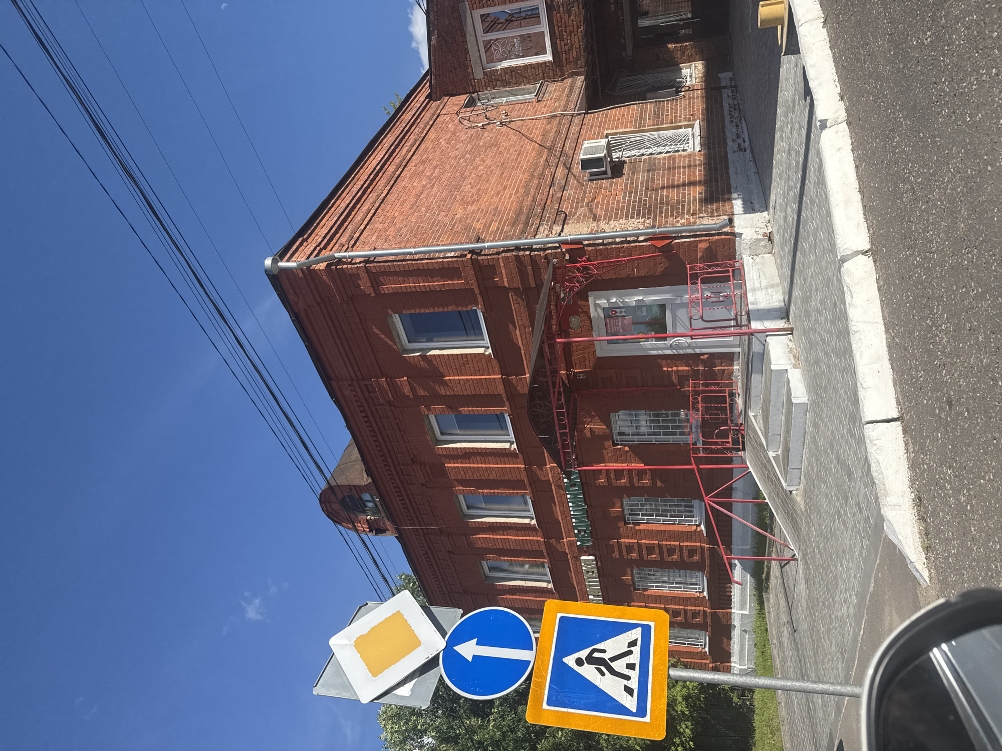 In the city of Vereya, Russia, a brick building with a red facade stands under a bright blue sky. The building, which appears to be a commercial establishment with the text "АПТЕКА" (Pharmacy) visible above the entrance, features multiple windows with metal grates and a red awning over the doorway. A ramp and stairs lead up to the entrance, bordered by a red metal railing.
In the foreground, a traffic signpost displays three signs: a diamond-shaped sign with a yellow center and white border, a circular blue sign with a white upward-pointing arrow, and a square blue and orange sign with a white pedestrian crossing symbol. The ground is paved with gray interlocking bricks, and a portion of a car's side mirror is visible in the bottom left corner. Overhead, black electrical wires are strung across the sky. The scene is bathed in sunlight, casting shadows on the pavement.