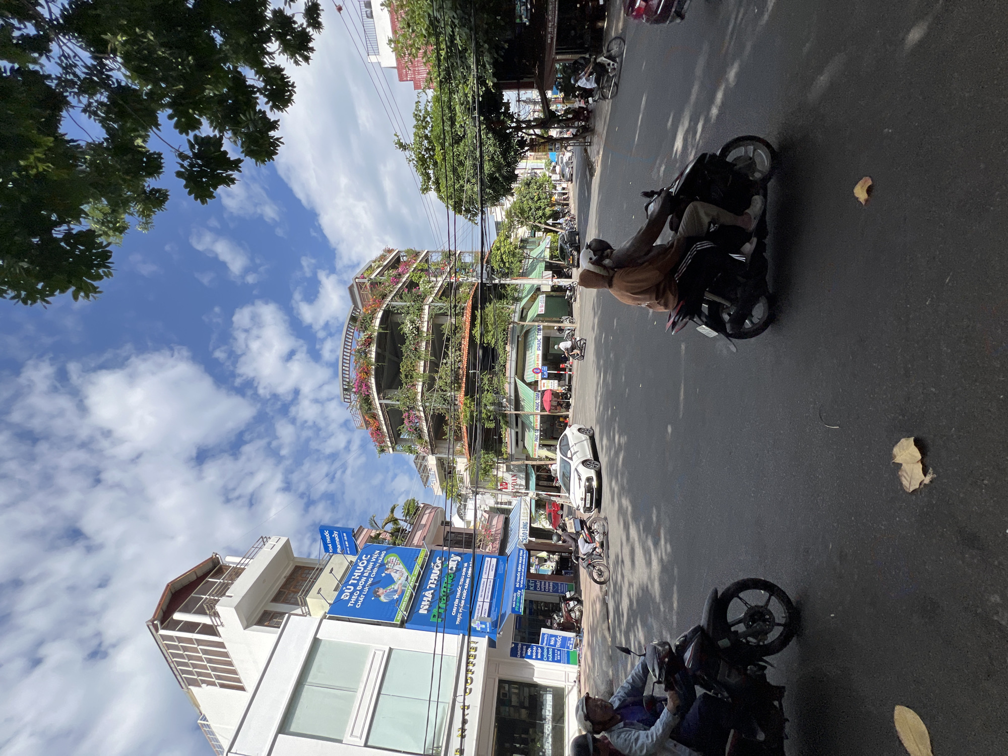 This is a street-level, eye-level shot on a sunny day in Nha Trang, Vietnam, likely depicting a commercial district. The foreground is dominated by an asphalt road with two motorbikes in motion. On the left, a motorbike is ridden by a person in a blue denim jacket and a helmet, accompanied by a passenger. On the right, another motorbike is ridden by a person wearing a brown hooded sweatshirt and light-colored pants, with a passenger also wearing a helmet. Both motorbikes are moving forward.
In the midground, several buildings line the street. A white sedan is parked on the right side of the road. A distinctive multi-story building with a curved facade is visible, adorned with numerous plants and flowers on its balconies and upper levels, creating a green, terraced appearance. Various shops and businesses are situated at street level, including one with a prominent blue sign displaying Vietnamese text and "Pharmacy". Other buildings show typical urban architecture with awnings and signs.
The background features a clear blue sky with scattered white clouds. Trees with green foliage are visible, casting shadows on the road. Electrical wires and poles are also part of the urban landscape. The overall scene conveys a bustling city environment with traffic and commercial activity under a bright sky.