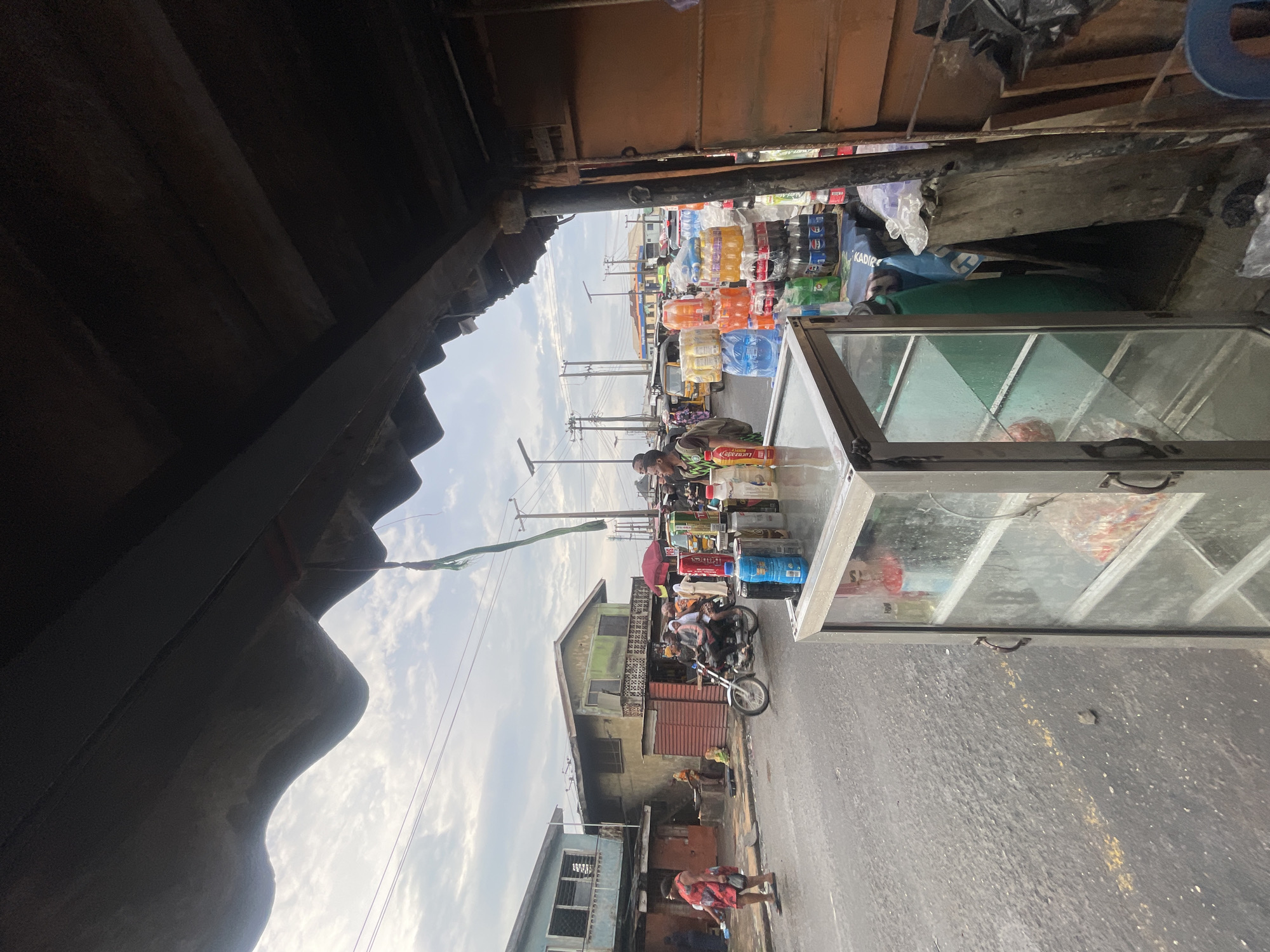 A street scene in Akowonjo, Nigeria, captures a bustling market environment under an overcast sky. The foreground is dominated by the underside of a weathered, corrugated metal roof, casting a shadow over a glass display case filled with various items. To the right, a wooden structure with an orange metal door stands, partially obscuring stacks of bottled drinks, including Coca-Cola and other sodas, alongside plastic containers and bags. A blue chair is visible to the far right.
The street in front of the market is paved and wet, suggesting recent rain. A yellow line is painted on the road. In the mid-ground, a motorcycle with three occupants—a driver and two passengers—travels from left to right. People are visible walking and standing along the street, with some gathered near stalls. Buildings line the street, appearing to be two-story structures with balconies and visible windows.
Further down the street, more stalls and a variety of vehicles, including a yellow minibus and other cars, indicate ongoing commercial activity. Power lines and utility poles stretch across the horizon. A green ribbon hangs from the roof in the upper right portion of the image. The overall impression is one of everyday life and commerce in a Nigerian urban setting.
