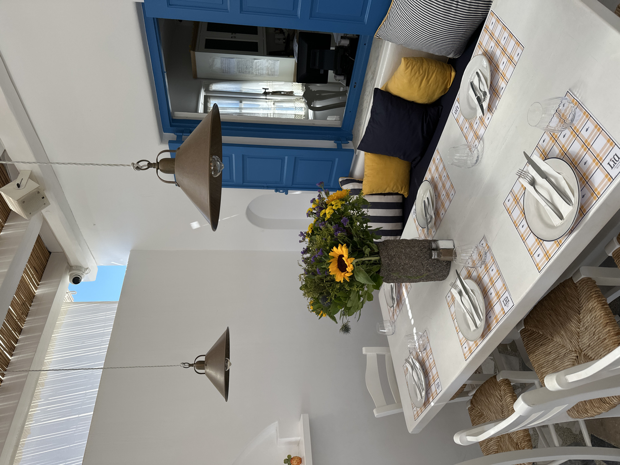 The image depicts a well-lit outdoor seating area at a restaurant in Ano Mera, Greece. A white rectangular table is set with white plates, cutlery, and glasses, each place setting resting on a yellow and white checkered placemat. In the center of the table, a rustic vase holds a large bouquet of sunflowers and other wildflowers. Behind the table, a blue shuttered window offers a glimpse into an interior space with a computer monitor. A built-in bench with dark blue and yellow cushions lines the wall beneath the window. Hanging pendant lamps with metal shades illuminate the scene, and sunlight streams through a slatted ceiling structure. White walls and chairs contribute to the bright, airy ambiance. A security camera is visible on the ceiling.