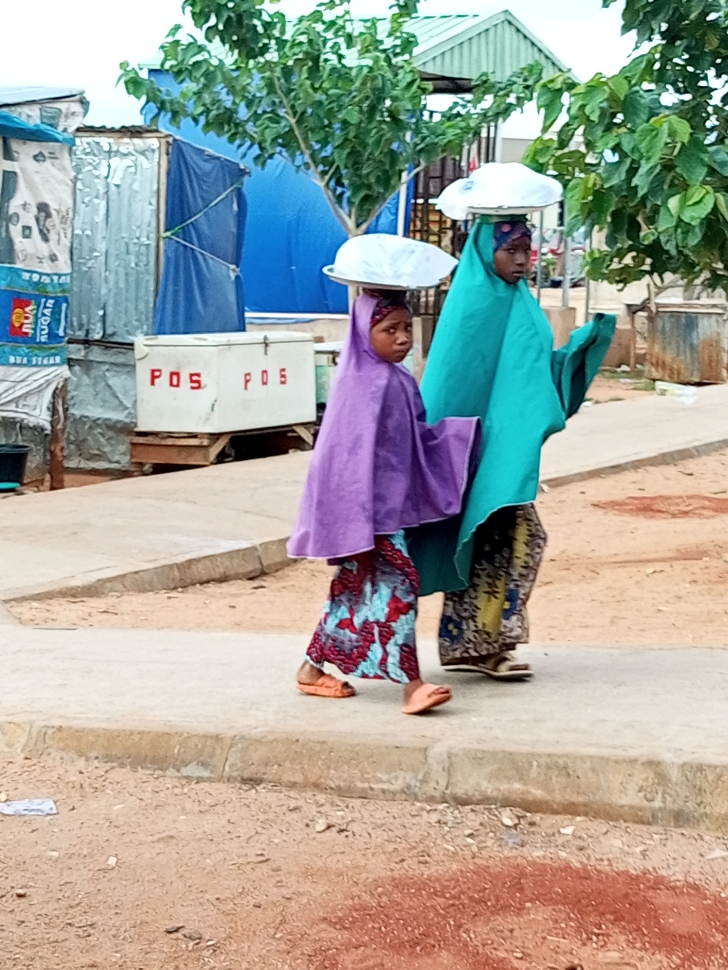 Two young girls are walking on a paved walkway in an outdoor environment. The girl on the left is wearing a purple headscarf and a colorful patterned dress. She is carrying a white disc-shaped object on her head. The girl on the right is wearing a teal headscarf and a patterned skirt. She is also carrying a white disc-shaped object on her head. Both girls appear to be walking in the same direction. In the background, there are various structures including corrugated metal buildings, a blue tarp, and a white box with red lettering that says "POS POS." There are also trees with green foliage visible. The ground is a mixture of dirt and sand, with some gravel. The overall scene suggests a marketplace or a residential area in Gombe, Nigeria.