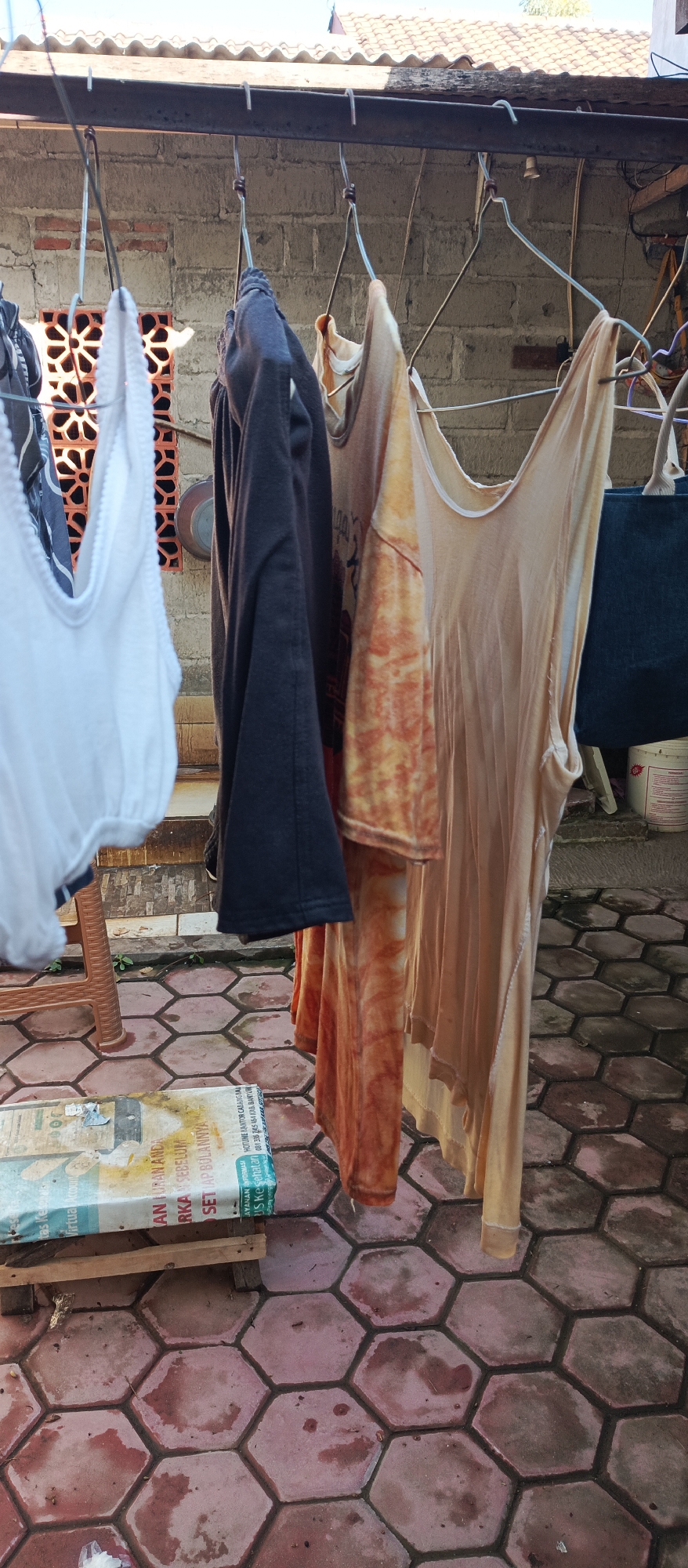 In Arjasa, Indonesia, a clothesline is hung with various garments, including a white bra, a dark t-shirt, a patterned t-shirt with orange and brown hues, and a light-colored tank top. The clothes are suspended from metal hangers. In the background, a brick wall with a decorative pattern is visible, along with a section of a tiled roof. The ground is paved with hexagonal tiles. A book or pamphlet lies on the ground in the foreground. The overall setting appears to be an outdoor area, possibly a backyard or a laundry drying space.