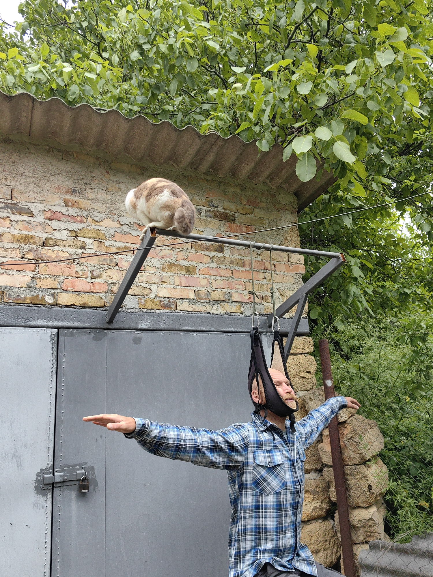 A medium shot shows a bald, middle-aged man standing in front of a gray garage door with his arms outstretched, and a cat sitting on a metal bar above the door. The man wears a blue plaid shirt and a black contraption attached to his head, possibly for traction. The garage door has a latch and a padlock. A brick wall rises above the door, topped with a corrugated metal roof. A cat with white and brown fur sits on the metal bar attached to the wall. Beyond the wall, lush green foliage fills the background. A rusted metal pipe stands next to the man, bordering a wall of stacked rocks. The location is an unknown city in the unknown region.