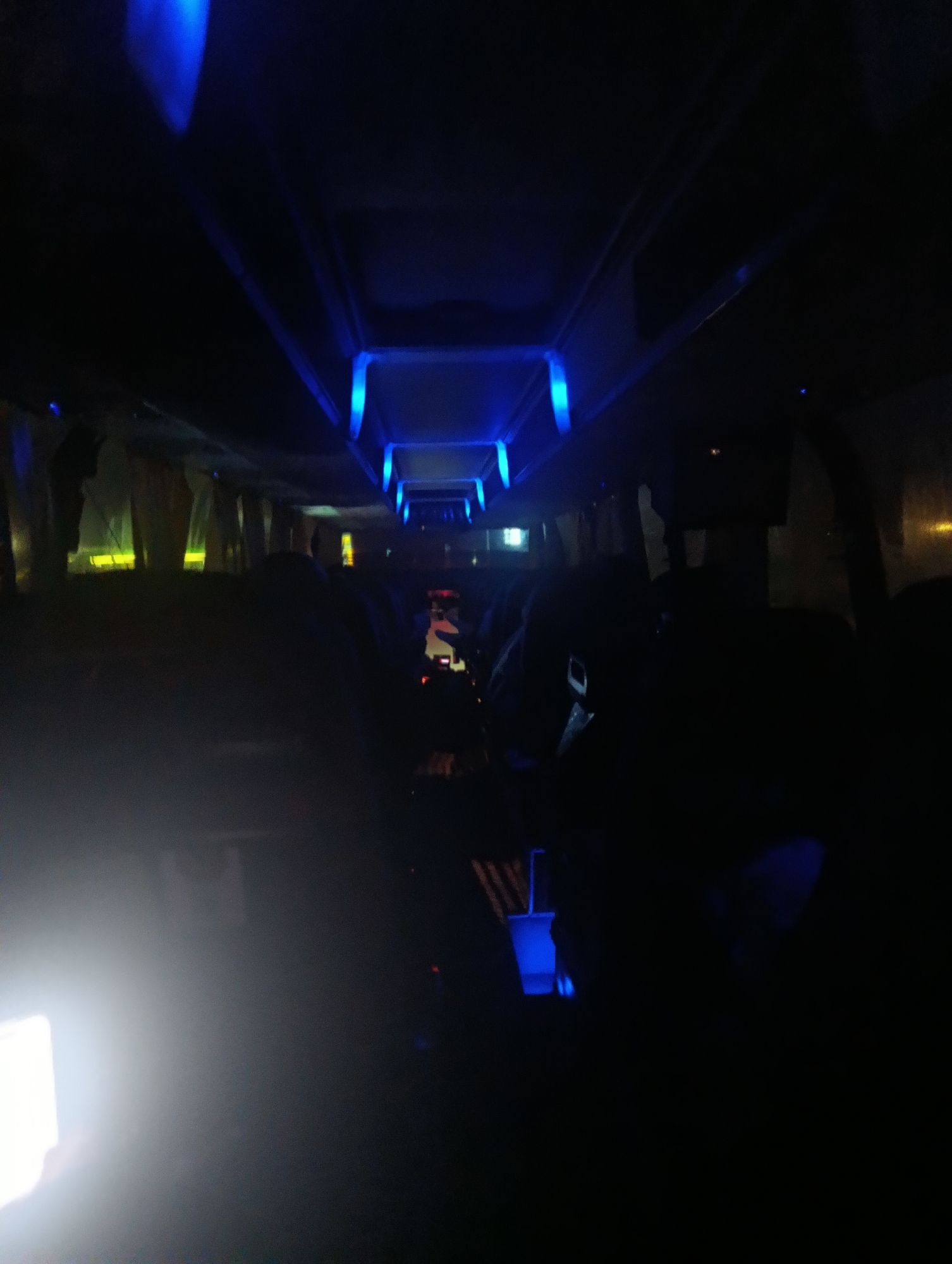 The image shows the interior of a vehicle, possibly a bus or train, illuminated by blue LED lights. The lighting is dim, creating a dark and atmospheric setting. In the foreground, on the left, a bright white light source is visible, possibly a camera flash or a phone screen. The interior appears to be empty of people in the immediate foreground, but silhouettes of passengers can be seen further back in the vehicle. The overhead compartments and lighting fixtures are visible. The image lacks any specific indicators of location within the city of Maiaky, Ukraine, or any identifiable landmarks. The scene is characterized by its low-light conditions and the presence of blue accent lighting.