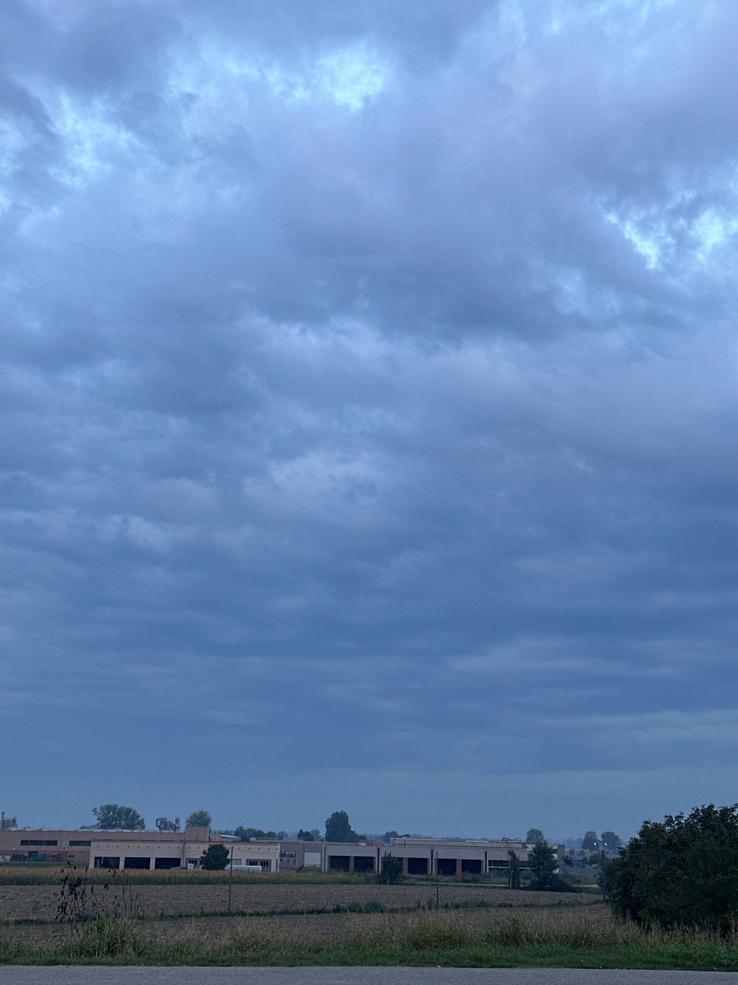 The image depicts a cloudy sky over a rural landscape near Dosolo, Italy. In the foreground, a dirt road runs horizontally across the bottom of the frame, bordered by dry grass and some sparse vegetation. Beyond the road, a field of dry crops stretches towards a row of industrial buildings in the middle ground. These buildings appear to be a complex of warehouses or factories, with a long, low profile and multiple sections. Some trees are scattered among the buildings. The sky is filled with heavy, dark gray clouds, suggesting an overcast or impending rainy day. The overall impression is one of a quiet, possibly industrial or agricultural area under a somber sky. There are no people or animals visible in the image.