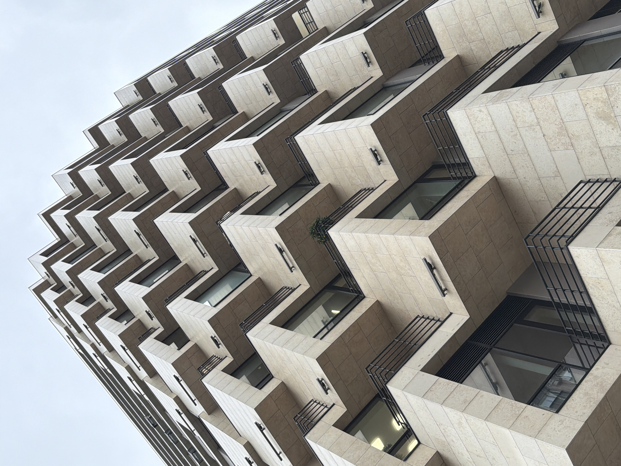 View of a tall building in Moscow, Russia, with balconies. The building is made of light-colored stone, and the balconies are arranged in a zigzag pattern. Black metal railings surround the balconies. Windows appear recessed into the building's facade. The sky is overcast.