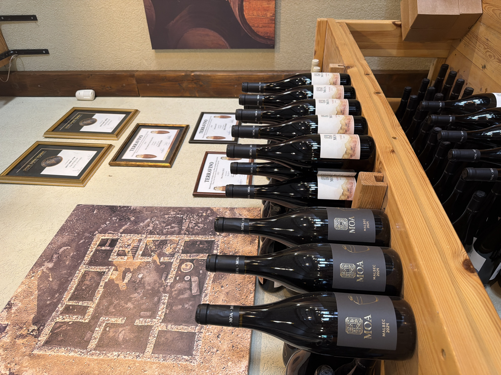 The image depicts the interior of a winery located in the Arava Tichona Regional Council, Israel. Prominently displayed are rows of dark glass wine bottles with labels indicating "MOA" and "MALBEC 2024". The bottles are arranged on light-colored wooden shelving. Above the bottles, a large framed photograph or artwork displays an aerial view of an archaeological dig site with stone foundations. To the right of the artwork, framed certificates or awards are mounted on the wall. A thick wooden beam runs vertically along the right side of the frame. The overall environment suggests a retail or tasting area within a winery, with a focus on product display and possibly recognition of achievements. No people are visible in the image.