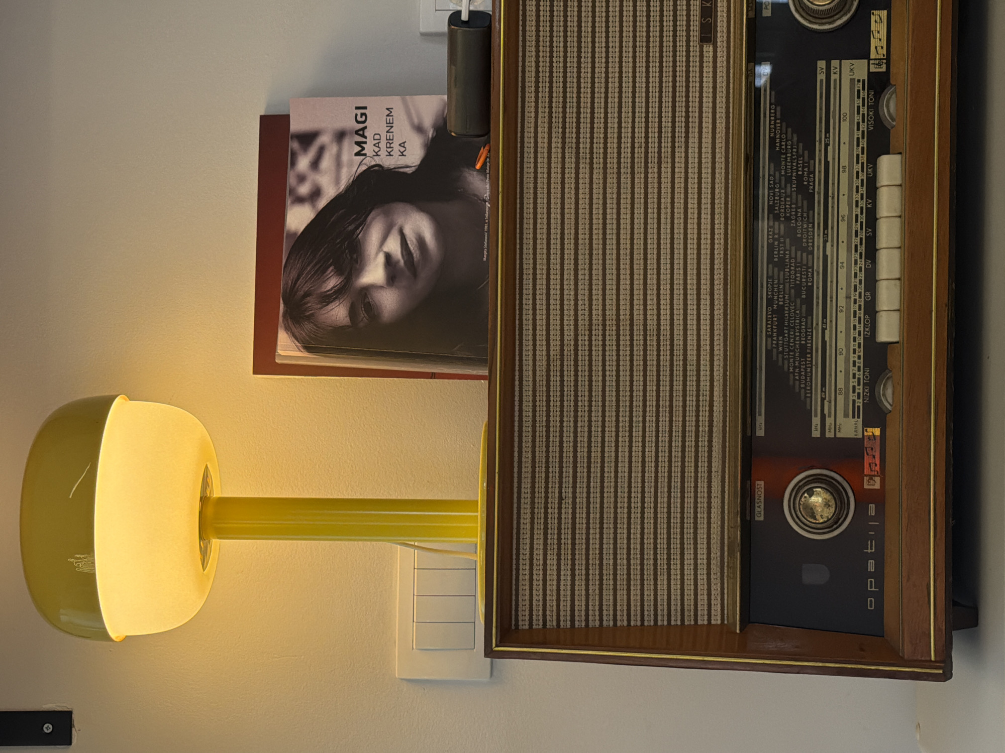 Here's a description of the image:
This indoor shot presents a partially visible vintage radio with a wood grain cabinet hanging on a wall. Next to the radio is a book titled "Magi Kad Krenem Ka," with a grayscale photo of a person on the cover. A yellow, spherical lamp with a cylindrical arm is attached to the wall below the book, near a multiple light switch panel. The radio features a speaker grille, station dial, and knobs. The light is illuminated, casting a warm glow on the surrounding wall. The location is within the city of Palilula Urban Municipality, Serbia, as evidenced by the station names listed on the radio.