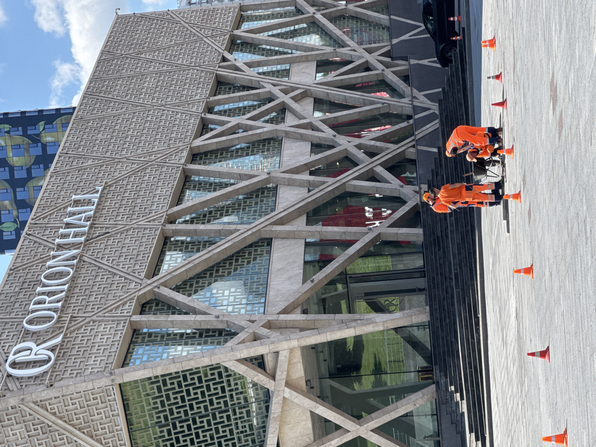 Four construction workers in bright orange high-visibility vests and pants are gathered on a paved plaza in front of a modern building. The building has a distinctive geometric facade with large glass windows and an "ORION HALL" sign in white lettering. The workers are interacting around a wheelbarrow, with one worker appearing to be directing others or examining something on the ground. Several orange traffic cones are scattered on the plaza, suggesting a work or construction area. A black car is parked to the right of the scene. The sky is partly cloudy. The image appears to be taken in an urban environment.