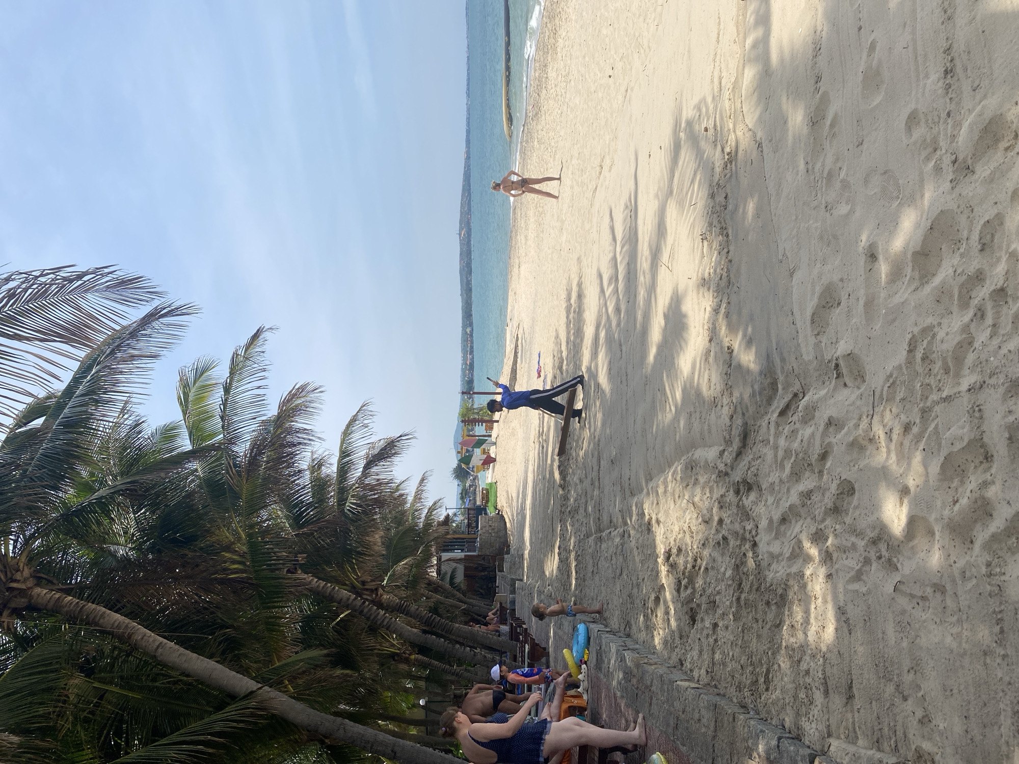 The image depicts a sunny day at a beach in Phu Thuy Ward, Vietnam, with the ocean visible in the background. Several people are present on the sandy shore. On the left side of the image, a woman in a dark blue swimsuit stands near a low stone wall, facing away from the camera. Next to her, a man in dark swimming trunks is partially visible. A young child in a blue and white striped swimsuit stands on the sand, facing the ocean. Further to the right, a person in a blue shirt and dark pants is walking away from the camera, holding a long wooden object. On the right side of the beach, a woman in a black bikini stands with her hands on her hips, facing the ocean. The beach is lined with palm trees on the left, casting shadows on the sand. Beach chairs and loungers are set up near the stone wall. The water appears calm with a distant shoreline visible.