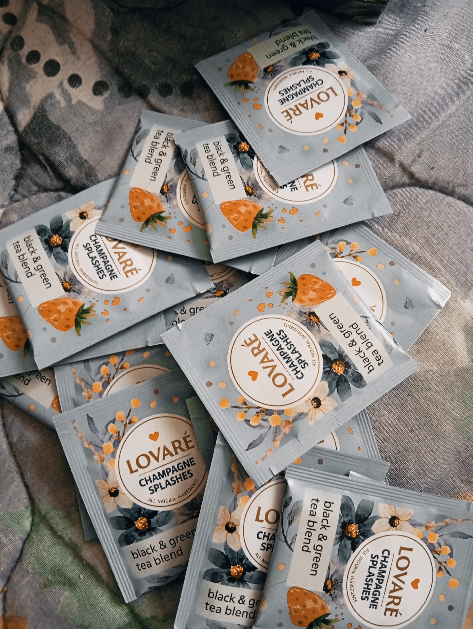 The image shows a collection of "LOVARÉ CHAMPAGNE SPLASHES" tea bags scattered on a surface with a patterned fabric. Each tea bag is light blue and features a picture of a strawberry, flowers, and the brand name. The text "black & green tea blend" and "ALL NATURAL INGREDIENTS" are also visible on the packaging. The location in the city of Poltava, Ukraine, is not determinable from the image content.
