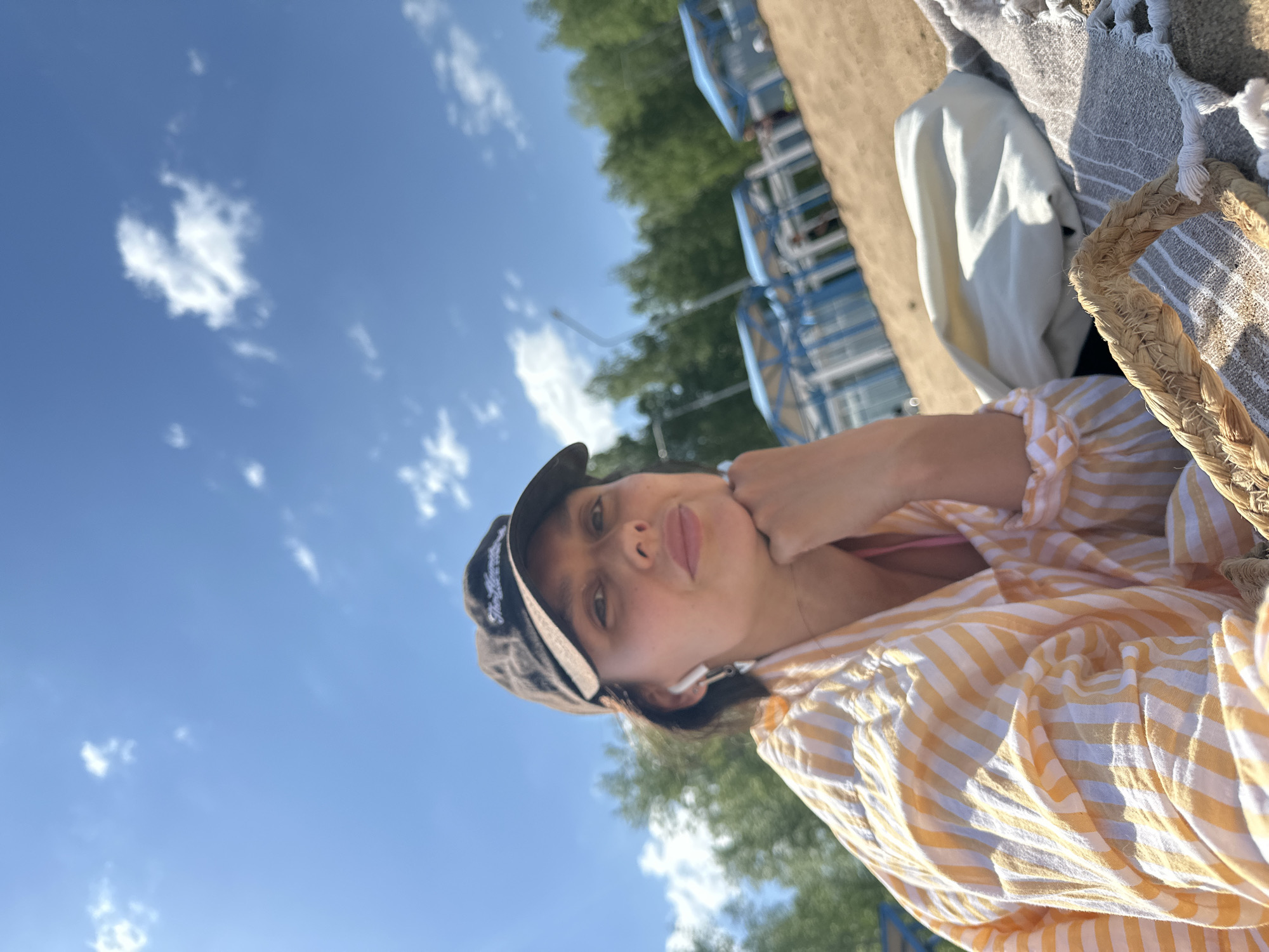 A woman wearing a black baseball cap and a yellow and white striped shirt is seen from a low angle against a sunny sky with scattered clouds. The woman's face is visible, and she is resting her chin on her hand. In the background, there are green trees and a blue metal structure, possibly a playground or park equipment. A beach bag and a towel are partially visible in the upper right corner. This scene could be taking place in Chaikovsky, Russia, potentially in a park or recreation area near a water body.