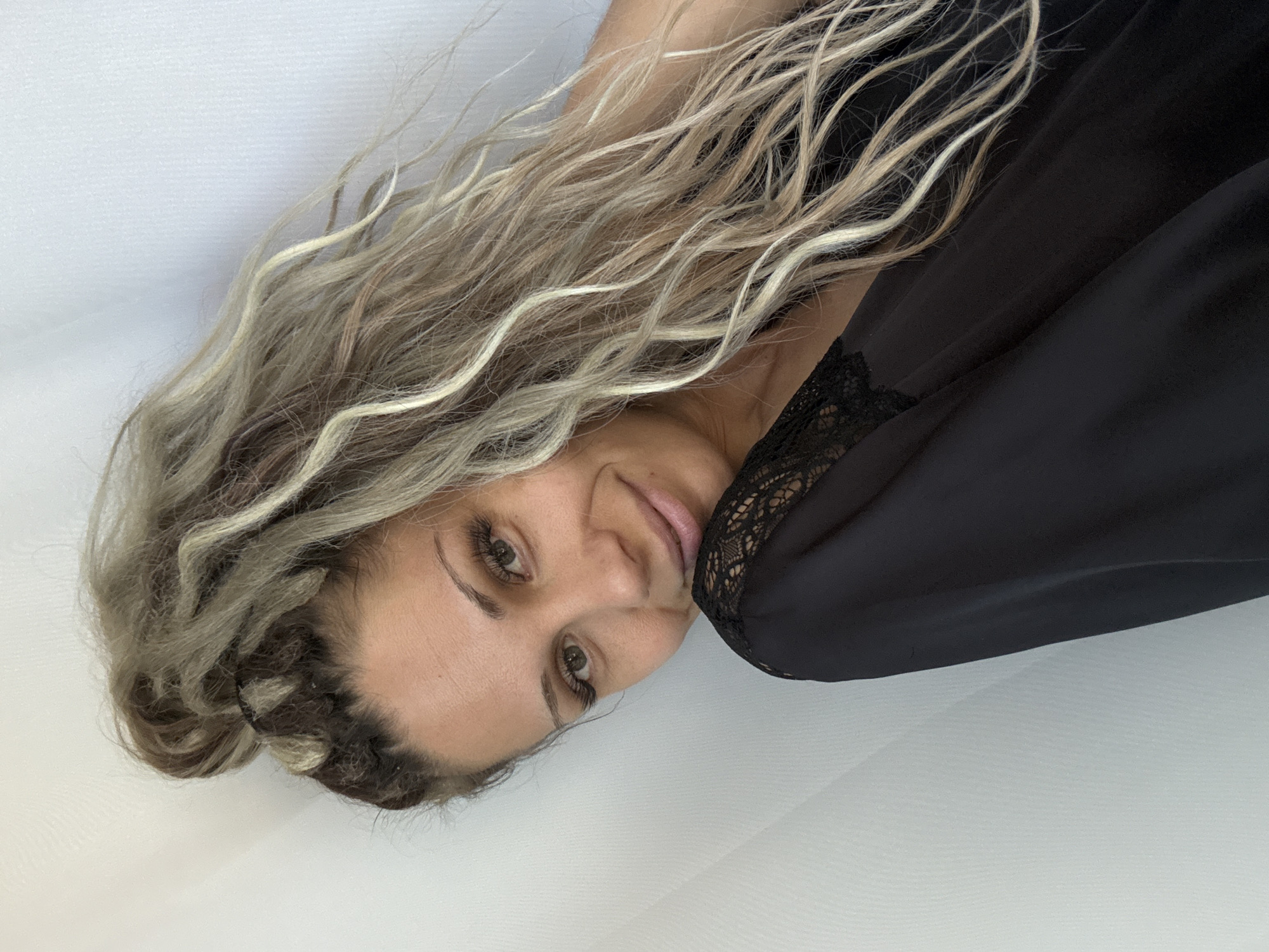 A young woman with blonde, multi-toned, dreadlocked hair is positioned in a selfie-style portrait. Her hair is styled in a high bun with strands cascading down. She is wearing a black garment with lace detailing on the shoulder. The background consists of a light-colored, textured surface, possibly a wall or curtain. The lighting suggests an indoor setting. The woman is looking towards the camera with a slight smile, her head tilted. There is no identifiable location in the city of Rusanovo, Russia present in the image.