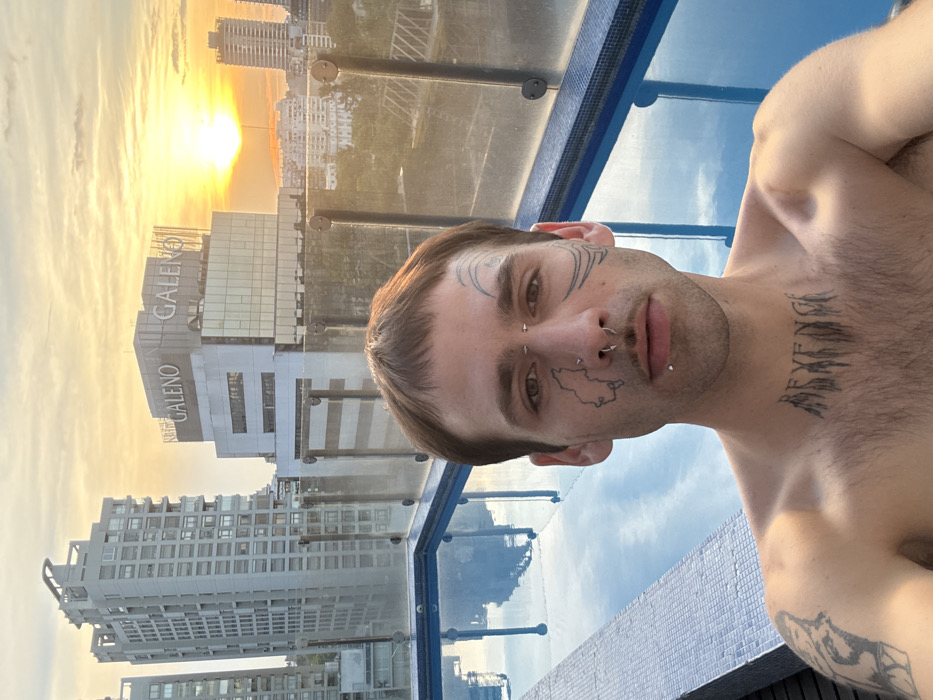 A shirtless person with face and neck tattoos poses against a backdrop of high-rise buildings and a sunset. Visible piercings are on the face. The person stands next to a swimming pool with a glass barrier, reflecting the sky and nearby buildings. Buildings with signage like "GALENO" can be seen.