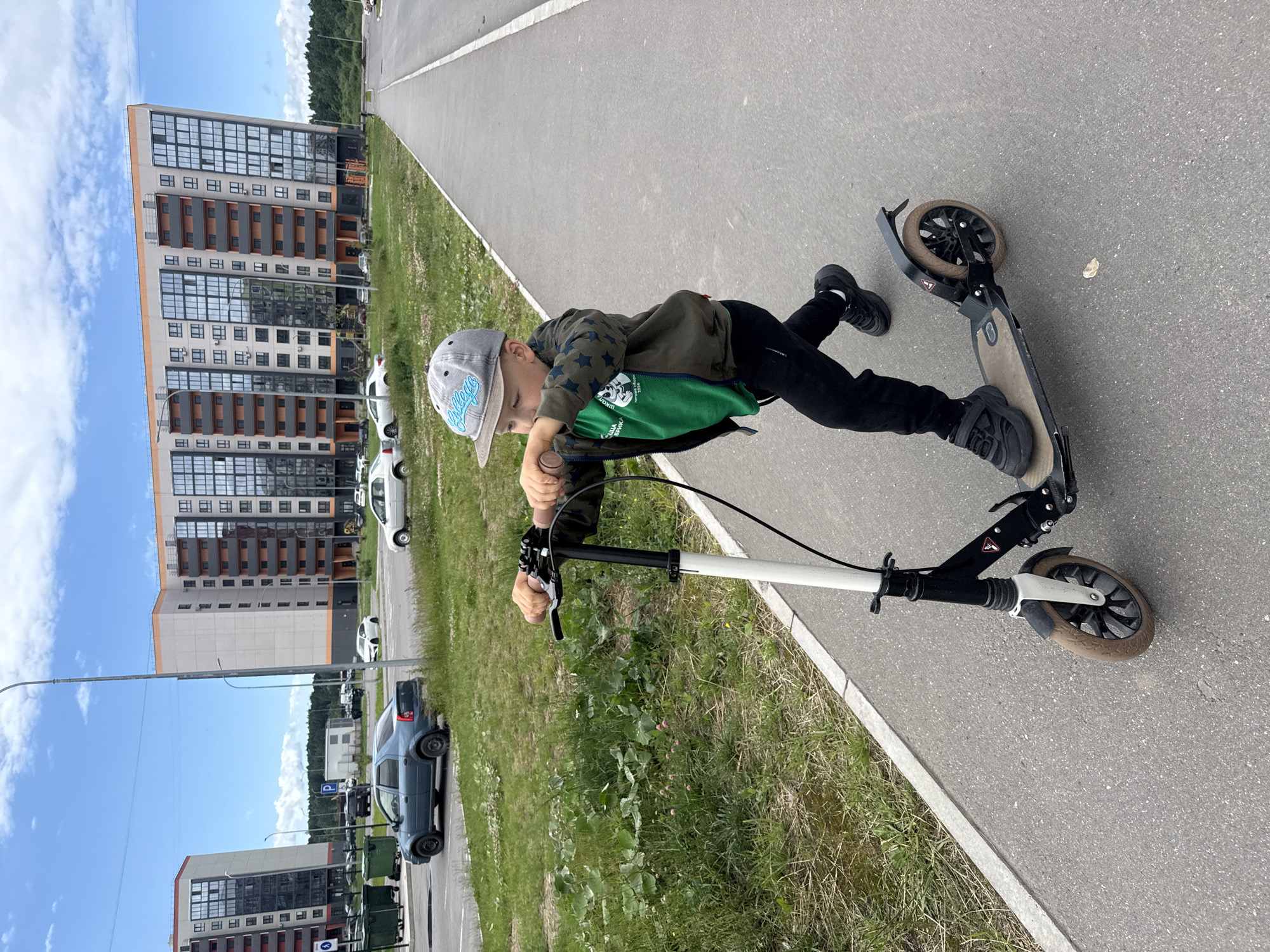 A young boy wearing a grey baseball cap, a green t-shirt with graphics, and a camouflage patterned jacket, is riding a black and white scooter on a paved path next to a grassy area. He is wearing black pants and black sneakers. In the background, there are multi-story residential buildings with modern architecture, a paved road with parked cars, and a clear sky with scattered clouds. The scene appears to be in a newly developed residential area.