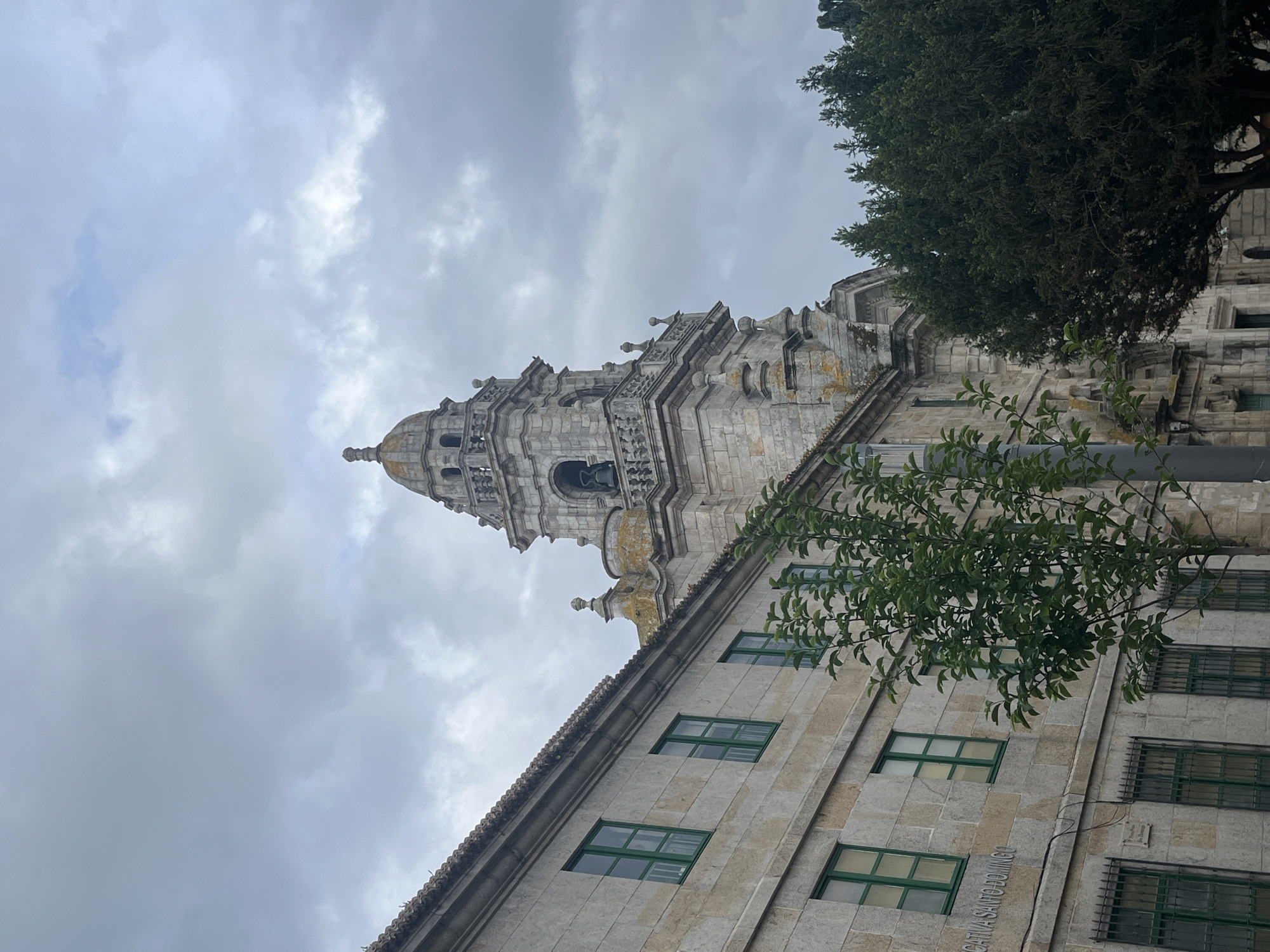 The image depicts a tall stone building with a tiered, ornate tower against a cloudy sky. The building has a tiled roof and multiple windows with green frames. A tree with green foliage is partially obscuring the building on the right side. A small plaque is visible on the building's facade, with faint inscription "CATHUA SANTO DOMINGO".
The scene suggests a location in A Coruña, Spain, specifically near the Santo Domingo Church.