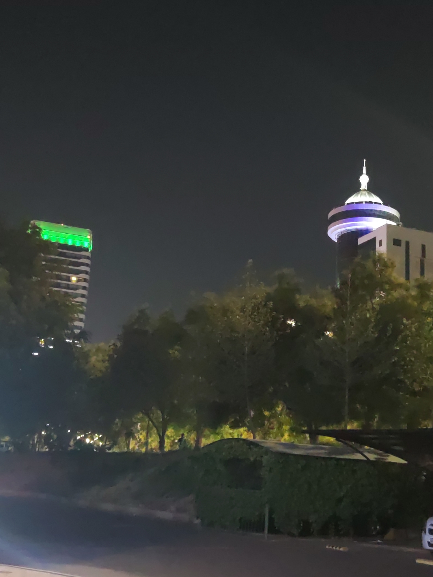 The image depicts a night scene in Tashkent, Uzbekistan, featuring two prominent buildings. On the left, a tall, multi-story building is illuminated with bright green lights along its upper floors. To the right, a distinctive tower-like structure is visible, topped with a dome that is lit with purple and white lights. The foreground shows a dark road or pathway with some trees and foliage. In the mid-ground, a line of trees partially obscures the view of the buildings. Further back, beyond the trees, there are indications of streetlights and possibly some people walking. A white vehicle is partially visible on the far right. The sky is dark, indicating nighttime.