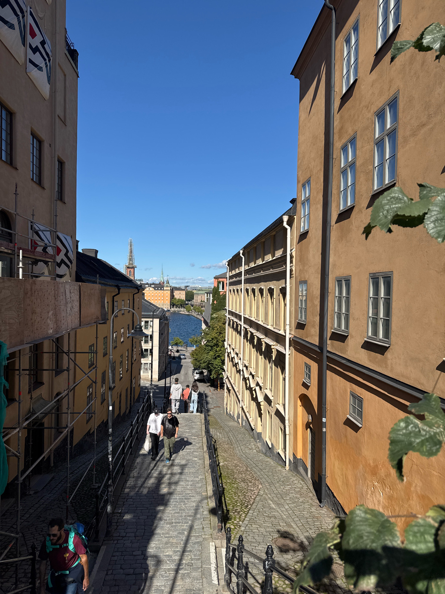 The image shows a cobblestone street in Gamla Stan, Stockholm, Sweden. The street slopes downwards towards a body of water, likely the Baltic Sea, with buildings on either side. The buildings are old, with typical Scandinavian architecture, featuring colorful facades and many windows. On the left side of the street, scaffolding and construction materials are visible, suggesting ongoing renovation work. Several people are walking down the street, some carrying bags, indicating it is a public space. The sky is clear and blue, and the sun is casting long shadows, suggesting it is a bright, sunny day. The overall impression is of a historic and picturesque urban environment.