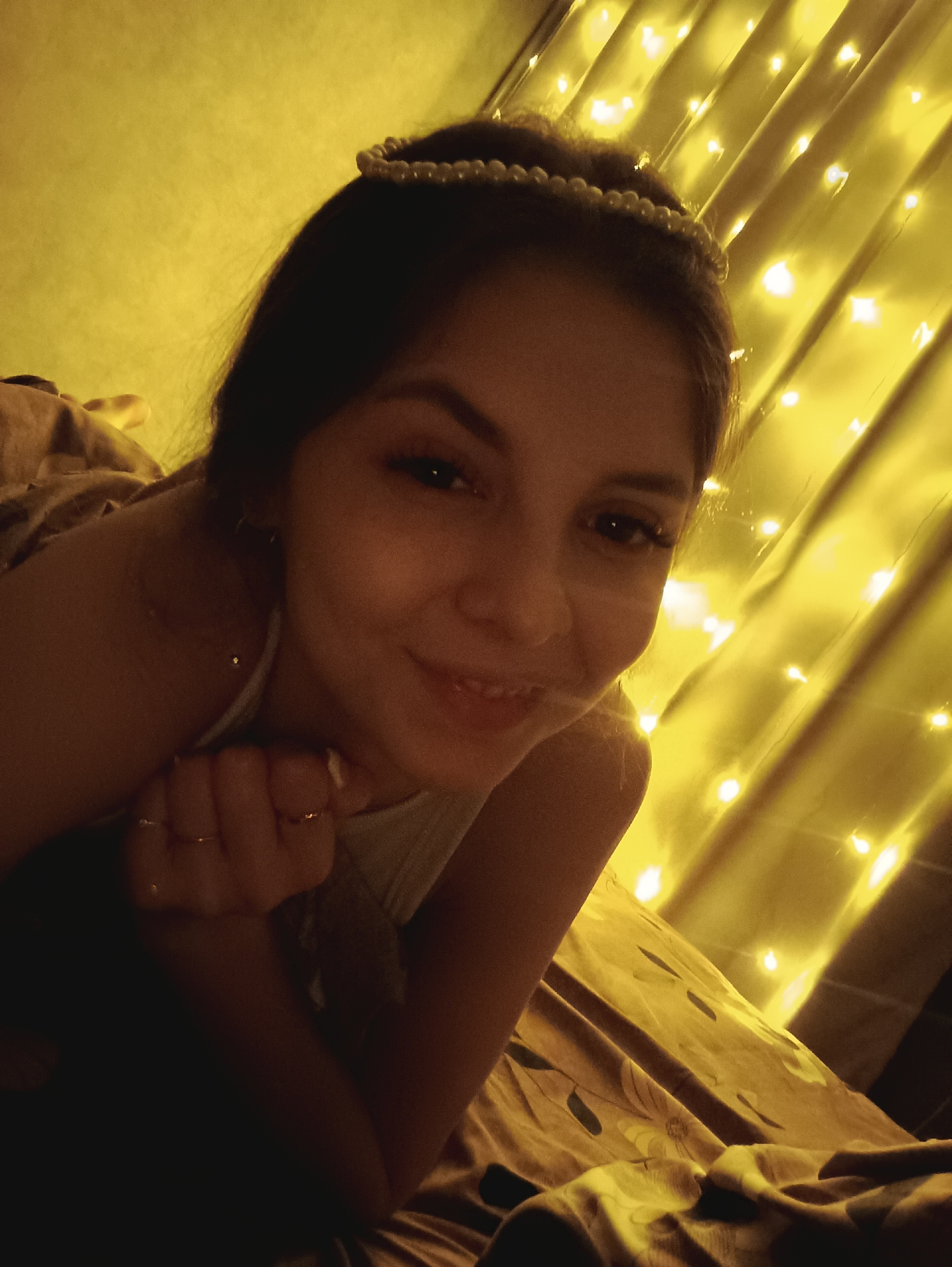 A young woman with dark hair and fair skin lies on a bed covered in a brown and gold floral patterned comforter. She is wearing a pearl-studded headband and a light-colored top. Her left hand is clenched near her face, adorned with rings on her fingers. The background consists of a wall covered in string lights, illuminating the scene with a warm, yellow glow. The location is unknown, there is no indication that the location is in the city of Chaikovsky, Russia.