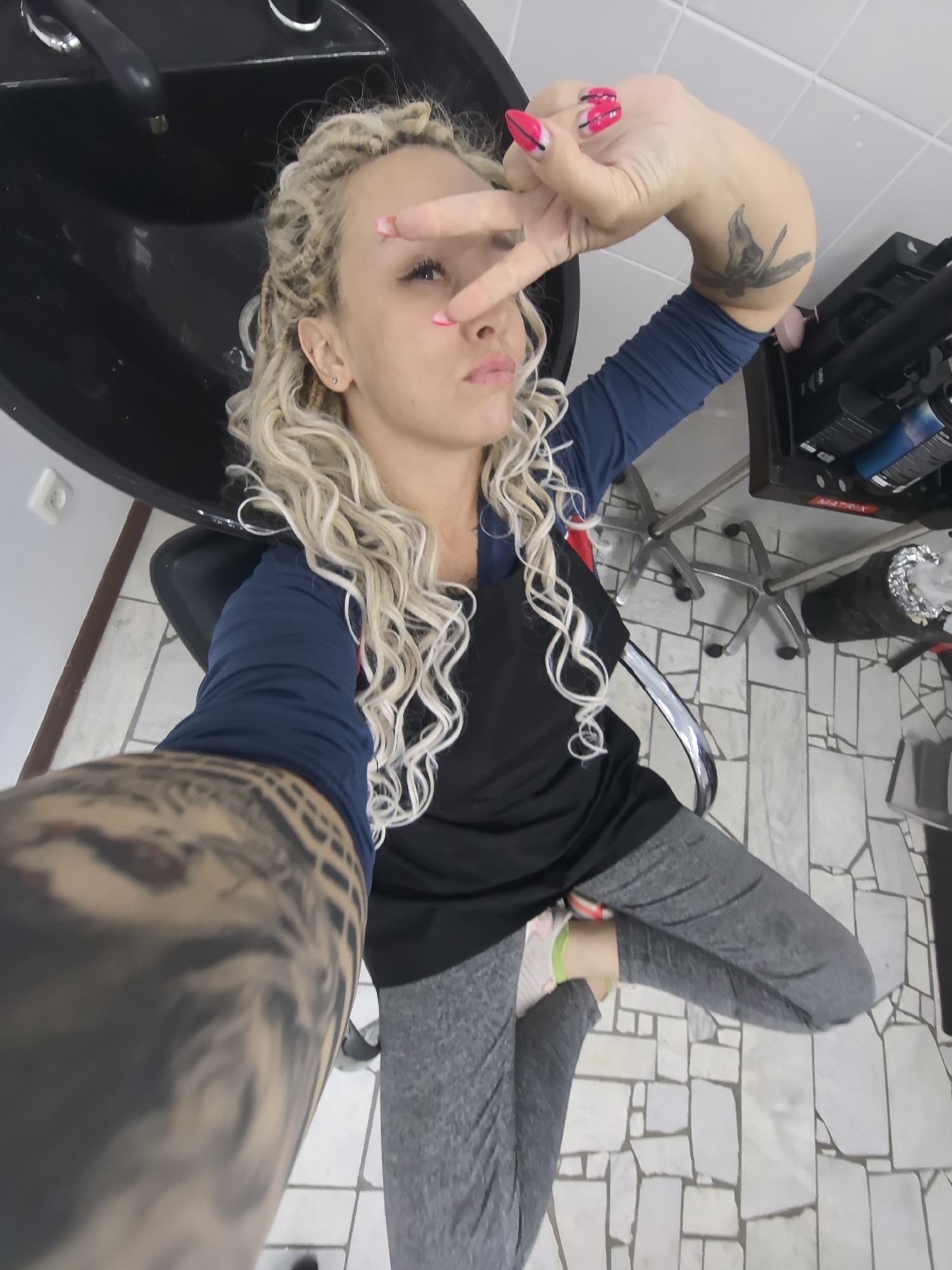 A young woman with blonde curly hair is taking a selfie in what appears to be a salon. She is sitting in a black salon chair, with a sink visible behind her. She is wearing a blue long-sleeved shirt and grey pants. She has a large tattoo on her left arm and a smaller tattoo on her right forearm. Her fingernails are painted bright pink and white. She is making a peace sign with her right hand, and her expression is playful. The floor is tiled with light-colored stones. In the background, there are salon chairs and equipment, including bottles of hair products. The lighting suggests it is daytime.