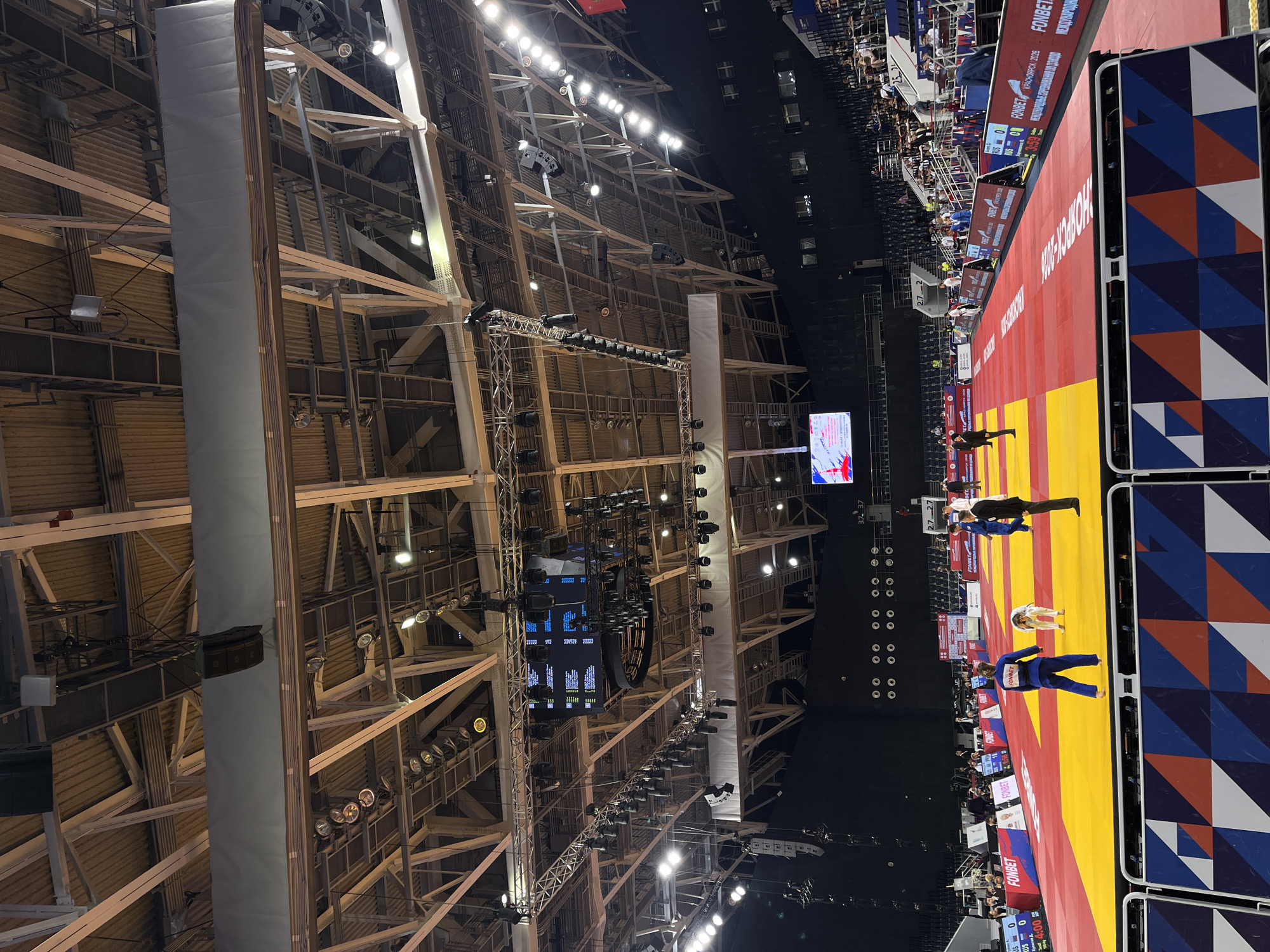 The image depicts an indoor judo competition taking place in the Platinum Arena in Krasnoyarsk, Russia. The arena features a bright yellow and red judo mat, surrounded by spectators in tiered seating. Overhead, a complex network of lighting rigs and sound equipment is suspended from the ceiling. Several individuals, dressed in judogi (judo uniforms) and officiating attire, are visible on and around the mat, engaged in the competition. Large screens display scores and event information. The overall environment is one of a professional sporting event with a focus on the judo competition.