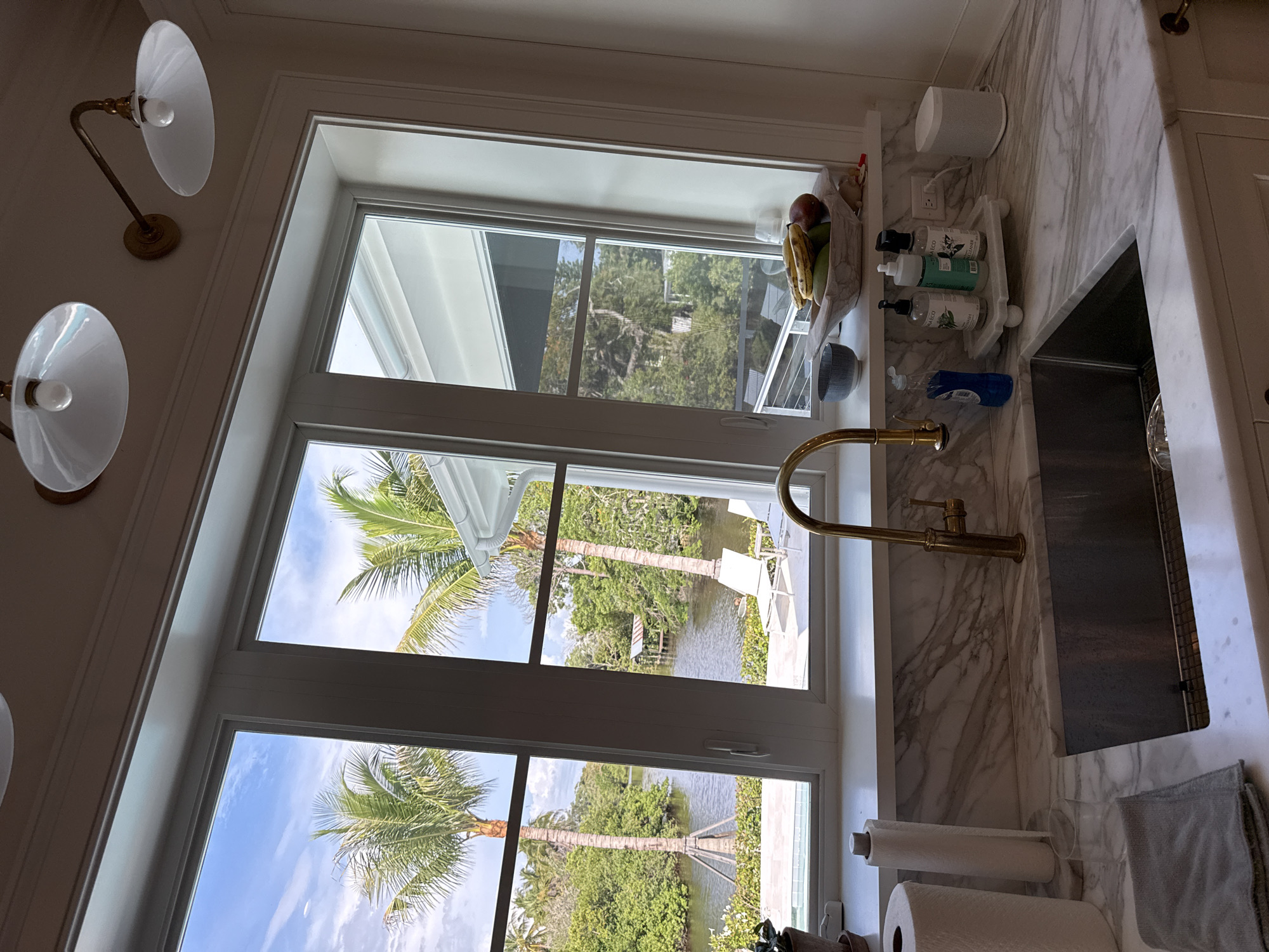 The image depicts an indoor view of a kitchen counter and sink area, looking out through a large window. The window offers a scenic view of a tropical waterway with palm trees and lush vegetation. In the foreground, the kitchen features a marble countertop with a stainless steel sink, a gold faucet, a roll of paper towels, a gray cloth, and several bottles of soap. A fruit bowl containing bananas, mangoes, and apples sits on a shelf above the sink. In the background, visible through the window, are two white lounge chairs and a small covered dock on the water. The ceiling above the window has two light fixtures. The scene appears to be in East Naples, Florida, United States of America, given the tropical vegetation.