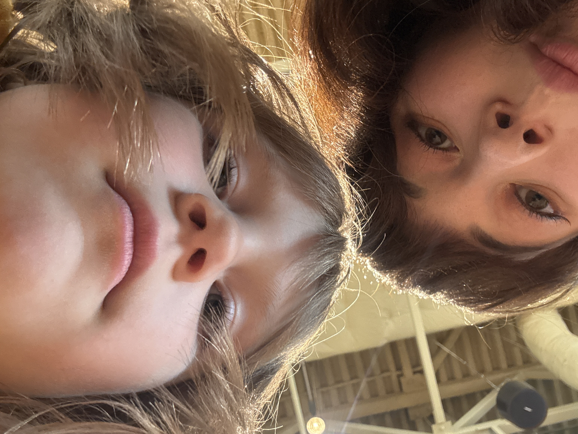 The image captures a close-up, low-angle portrait of two individuals, likely female, in an indoor setting located in the city of Perm, Russia. The subject positioned in the upper portion of the frame exhibits fair skin, light brown hair, and light eyes, with a slightly smiling mouth. The subject in the lower portion has moderately darker skin, dark brown hair, and dark eyes, looking directly towards the camera. Both individuals' hair shows significant highlights, consistent with strong overhead lighting. The blurred background reveals architectural details of a high ceiling, including white or light-toned structural beams and girders. Several light sources appear as diffused glows, and one distinct circular light fixture is visible in the upper-left background. A dark, cylindrical object, possibly an audio speaker, hangs from the ceiling structure in the lower-left background. No explicit actions or interactions beyond looking into the camera are present.