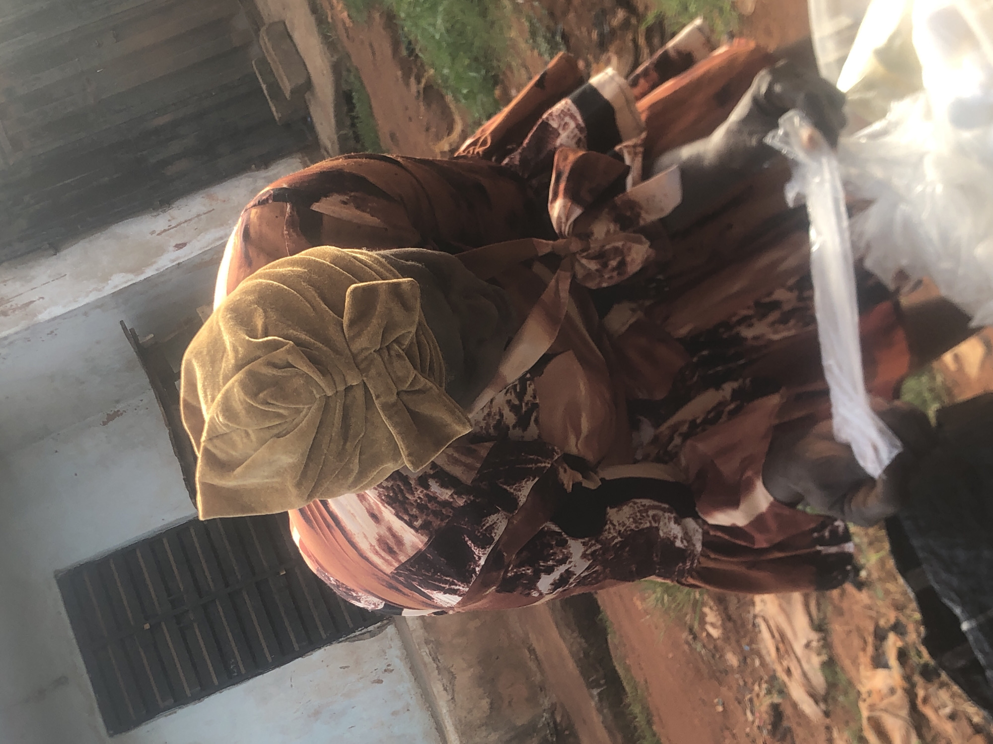 An adult female in Ukwu-Abwa, Nigeria, is outdoors, wearing a beige turban and a brown patterned dress. She is holding a plastic bag. The background includes a building with a window and an unpaved ground area.