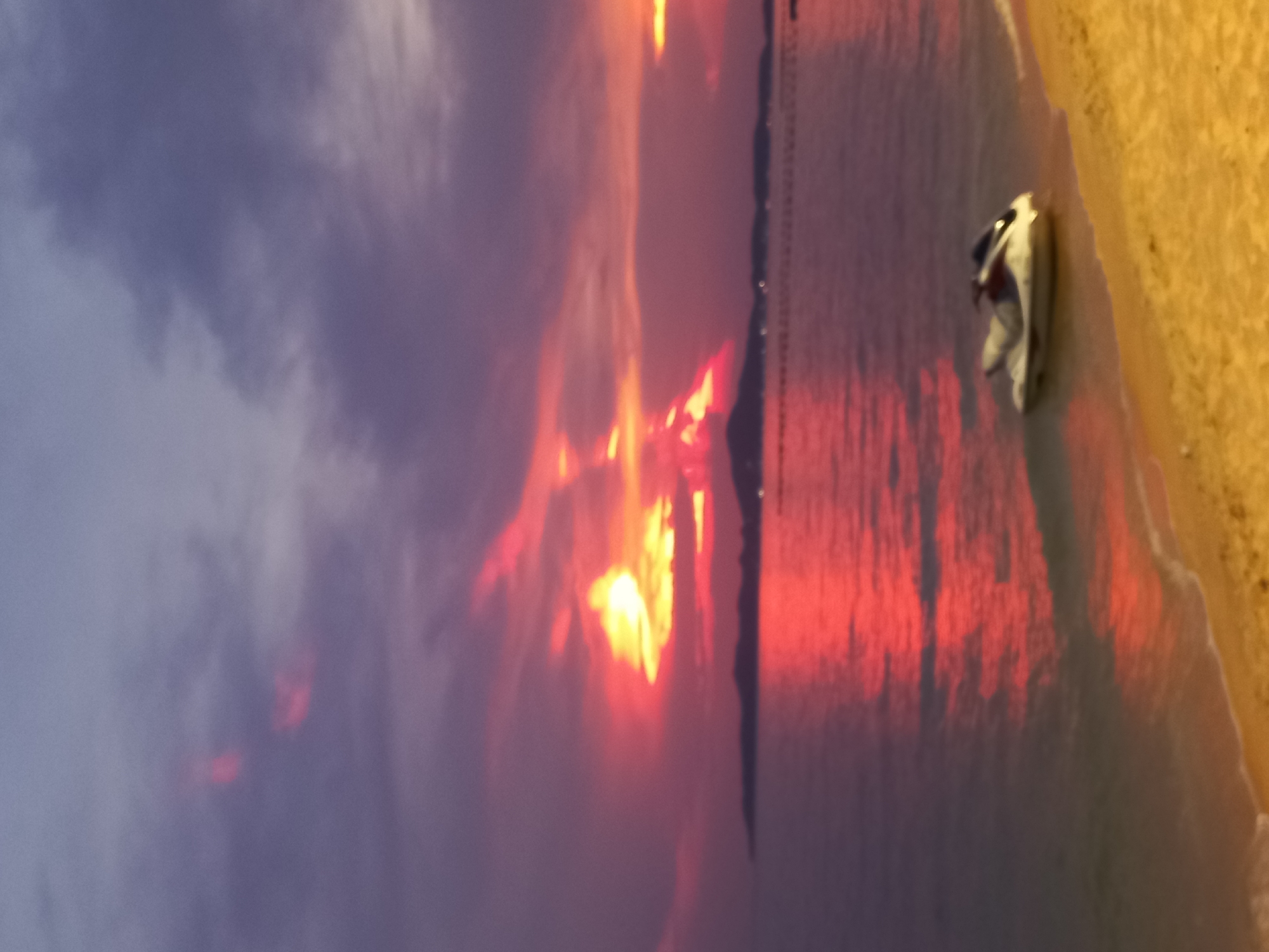 This sideways outdoor shot shows a beach at sunset in Pattaya City, Thailand. The sky is a mix of purple and gray clouds on the left and bright orange and yellow where the sun is setting. The sunset reflects off the water, creating a vibrant red hue. A beach is visible on the right side of the image, with a white object resting on it, likely a boat or kayak. There is a row of lights on the horizon.