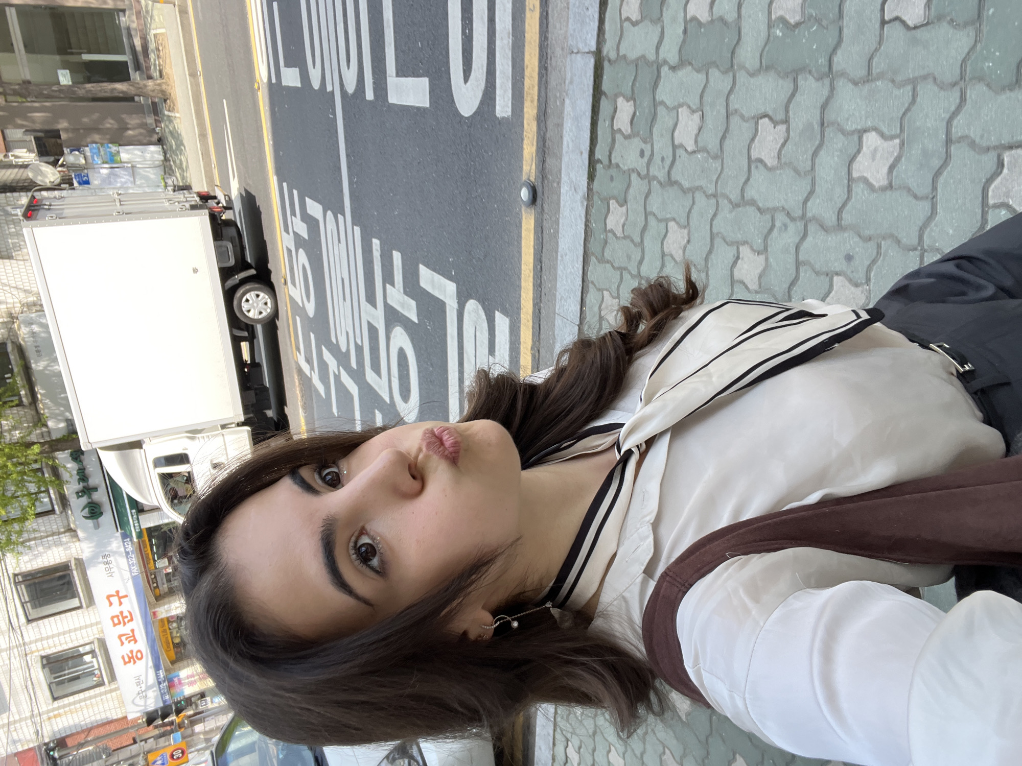 The image shows a young woman in a light-colored sailor-style top and dark pants, standing next to a street in Seoul, South Korea. She is taking a selfie and making a kissing face. The street has a truck on it and road markings in Korean. There are buildings in the background.