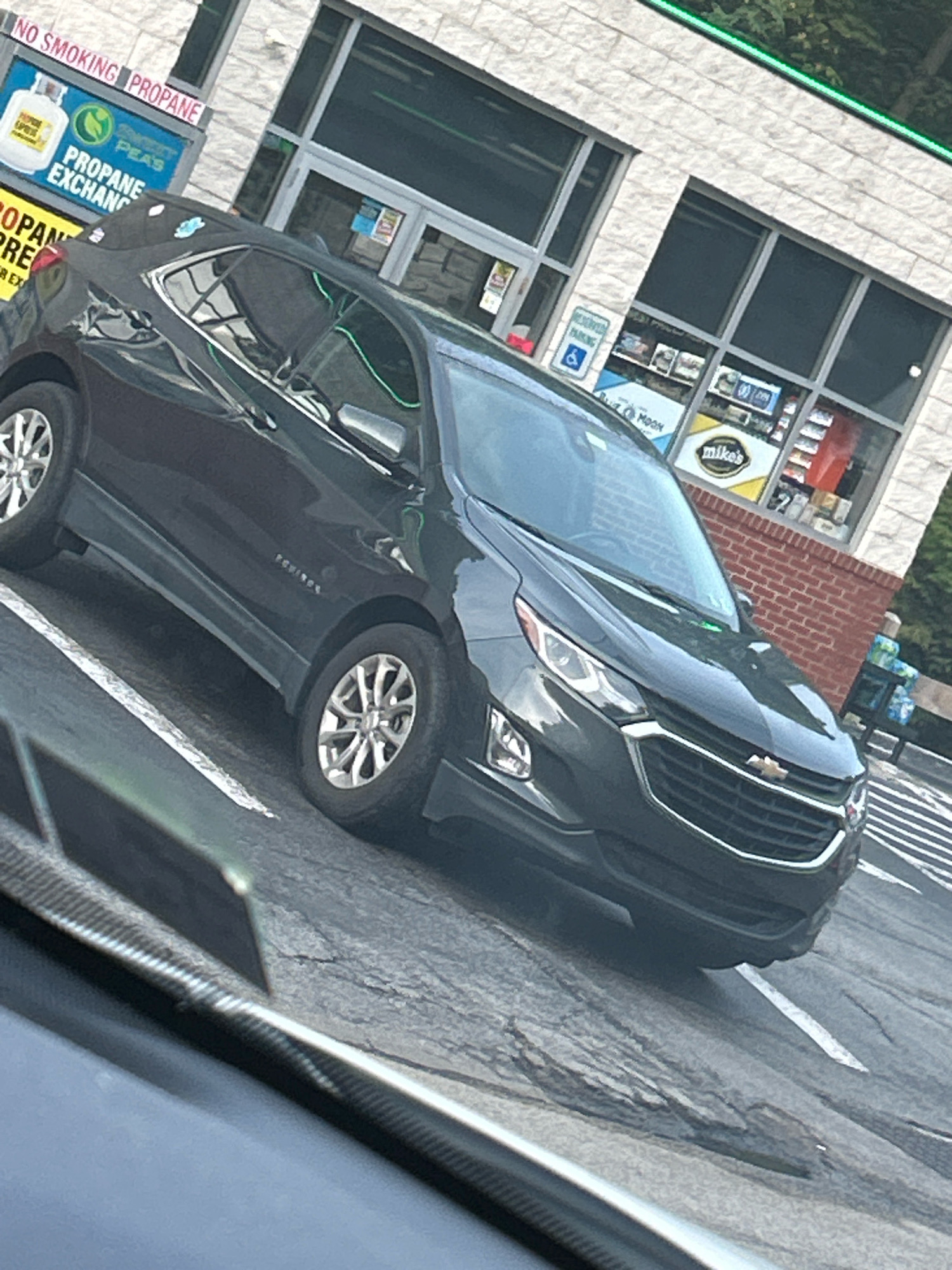 A dark gray Chevrolet Equinox is parked in a lot in front of a convenience store with a brick facade. The store has large windows displaying various products, including drinks and snacks. A sign above the entrance reads "NO SMOKING PROPANE". There is also a sign that says "PROPANE EXCHANGE" with a graphic of a propane tank. Another sign to the right indicates "mike's" with a yellow background. A blue and white sign with a wheelchair symbol designates reserved parking. The ground is asphalt with white parking lines. The car's headlights are on.
