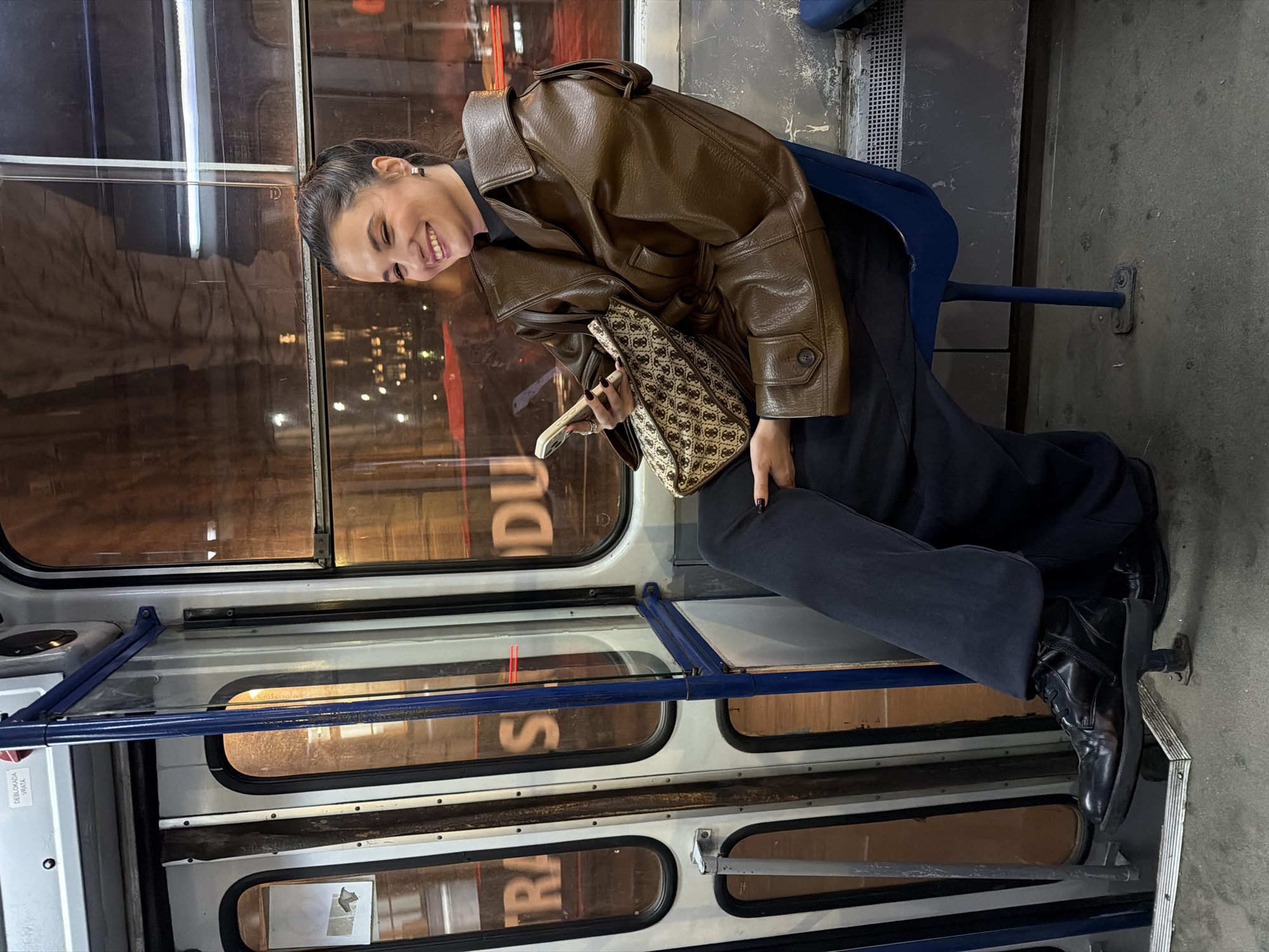 A female individual is seated on a blue plastic seat inside a public transit tram at night. She is positioned facing slightly to the right of the frame, smiling broadly. Her dark hair is pulled back, and she wears a brown, collared jacket over a black top, dark wide-leg trousers, and black boots. In her left hand, she holds a light-colored smartphone, and in her right hand, a patterned beige and brown handbag. Her fingernails are painted dark.
The tram's interior features narrow, vertically paneled doors with rounded windows and a blue metal handrail. A white sticker with the Cyrillic text "DEBLOKADA VRATA" (Door Unlocking) is visible on the upper interior wall, indicating the location is in Serbia, specifically within the city of Belgrade's tram system. Exterior city lights, including blurs of red and yellow, are reflected in the windows, along with partial reflections of signs displaying text fragments such as "TRAS" and "S DU." The floor is a dark, worn grey, and a metal ventilation grate runs along the bottom of the wall to the right of the subject.