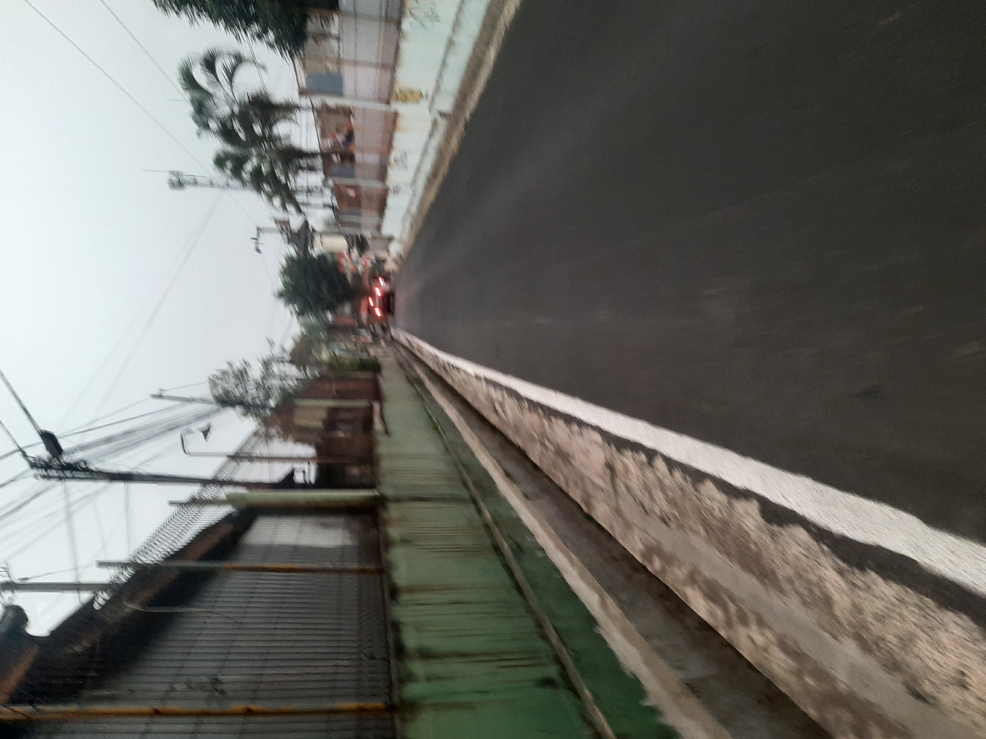 The image depicts a street scene in Purwanegara, Indonesia, characterized by a paved road with a white line marking the lane. On the left side of the road, there is a green wall with vertical streaks of rust or water damage. Above the wall, a metal fence is visible, followed by a building with a corrugated metal exterior. Utility poles and a complex network of electrical wires are prominent overhead. To the right of the road, there are buildings and trees, including palm trees. Two vehicles with their brake lights illuminated are traveling away from the camera in the distance. The sky is overcast, suggesting cloudy weather. The overall impression is of a somewhat industrial or commercial area.